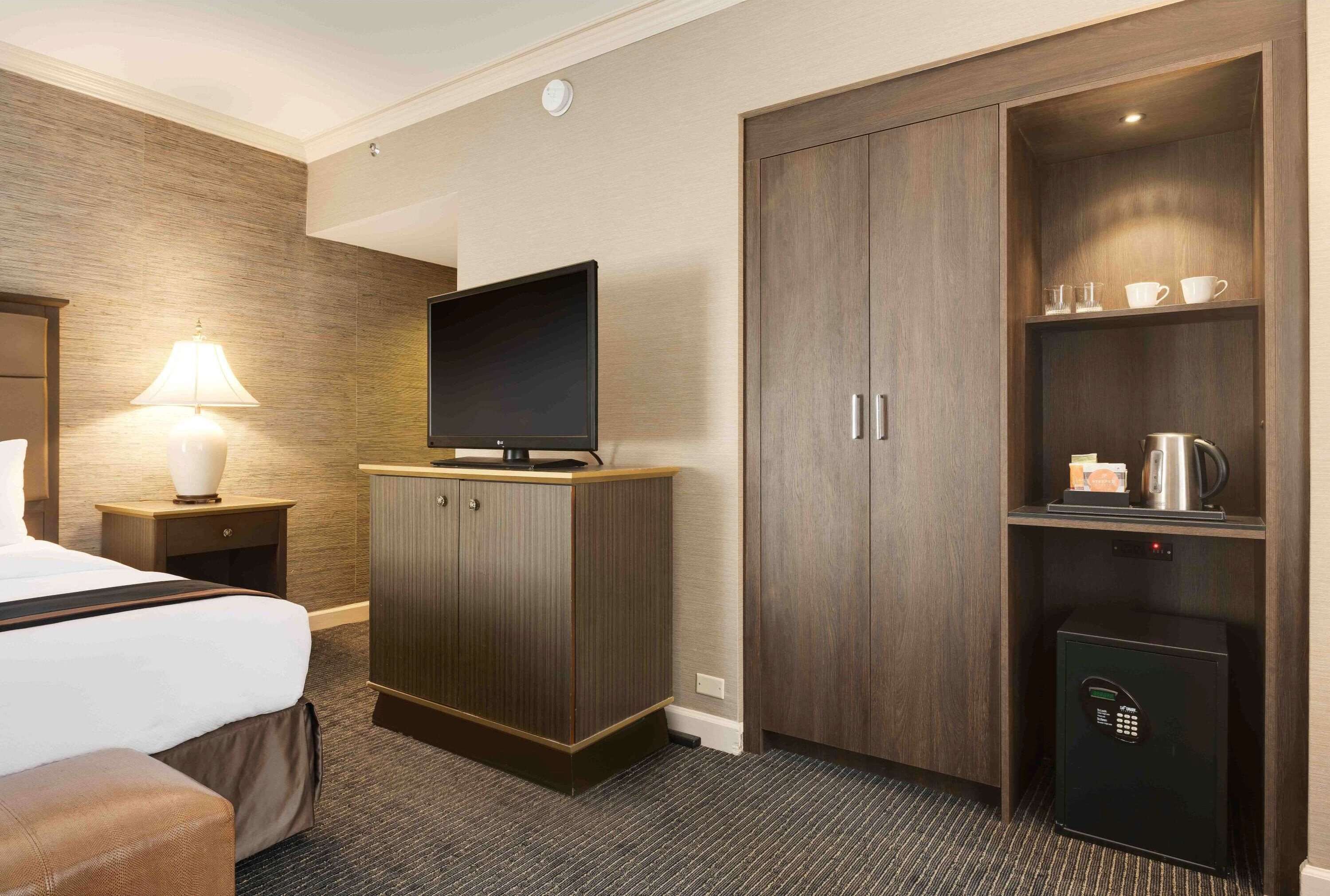 A hotel room with a bed, a TV on a wooden cabinet, a lamp, a minibar, and a safe.