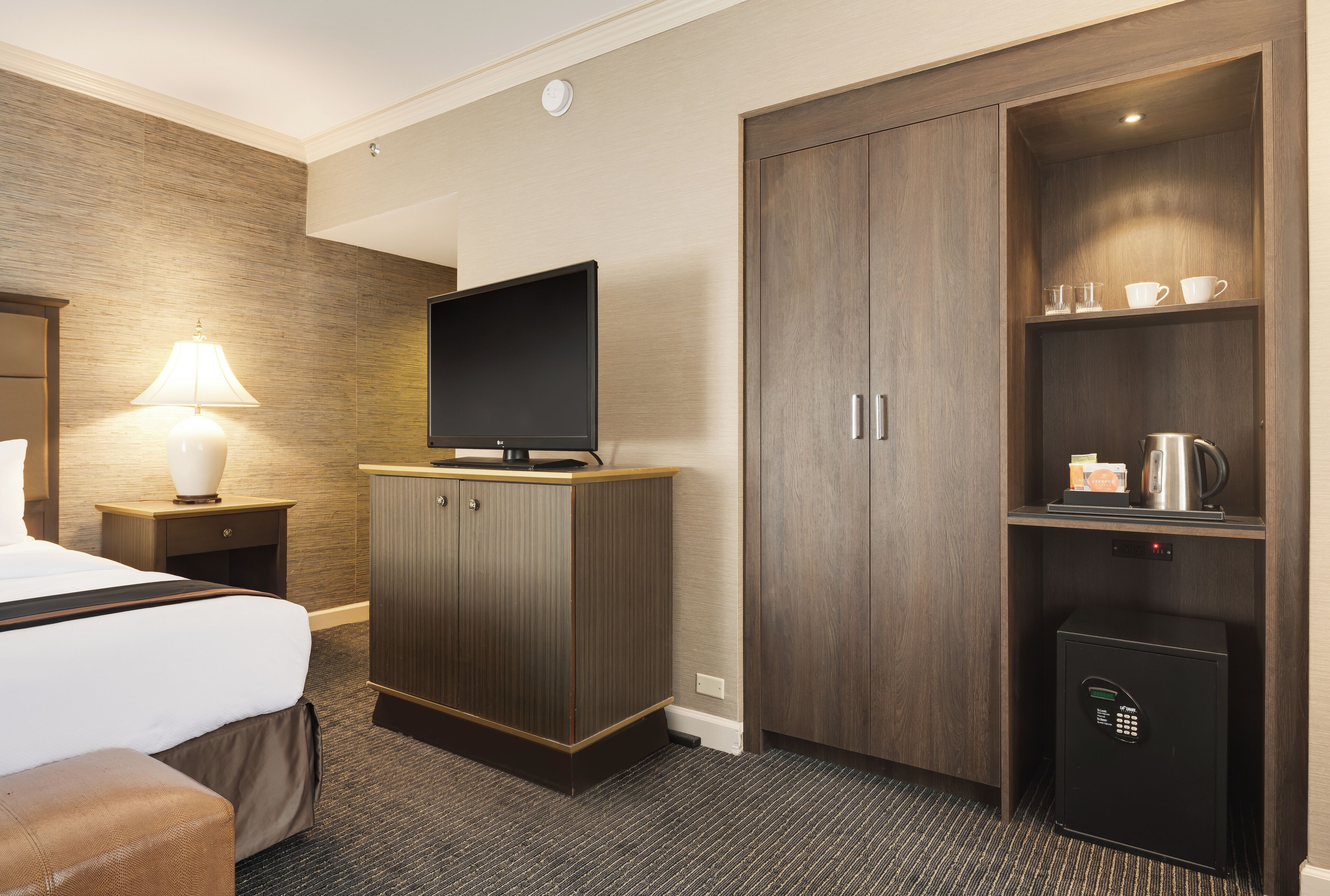A hotel room with a bed, a TV on a wooden cabinet, a lamp, a coffee maker, and a safe.