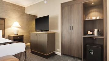 A hotel room with a bed, a TV on a wooden cabinet, a lamp, a coffee maker, and a safe.