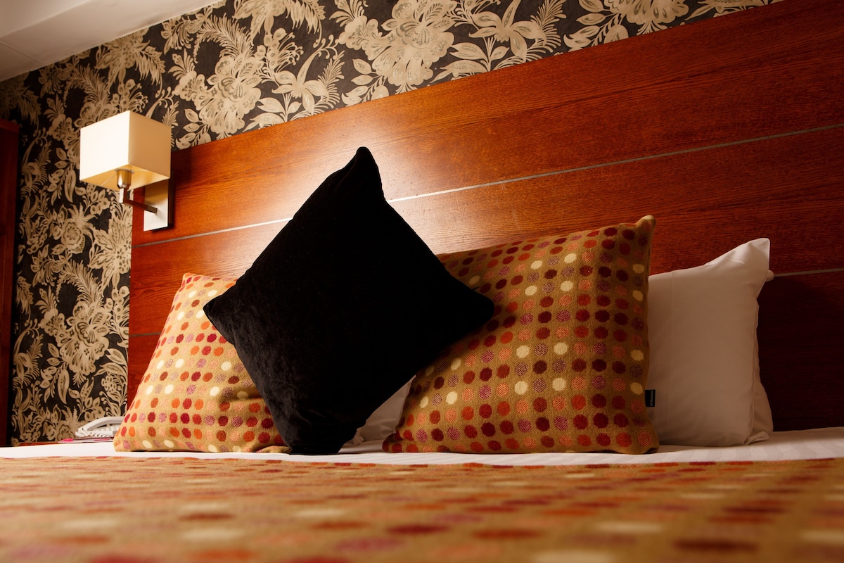 A hotel room with a wooden headboard, patterned wallpaper, and a bed with multiple pillows.