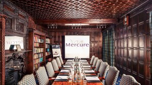 Un salón de conferencias con una larga mesa de madera, sillas estampadas, estanterías y una pantalla de proyector mostrando 
