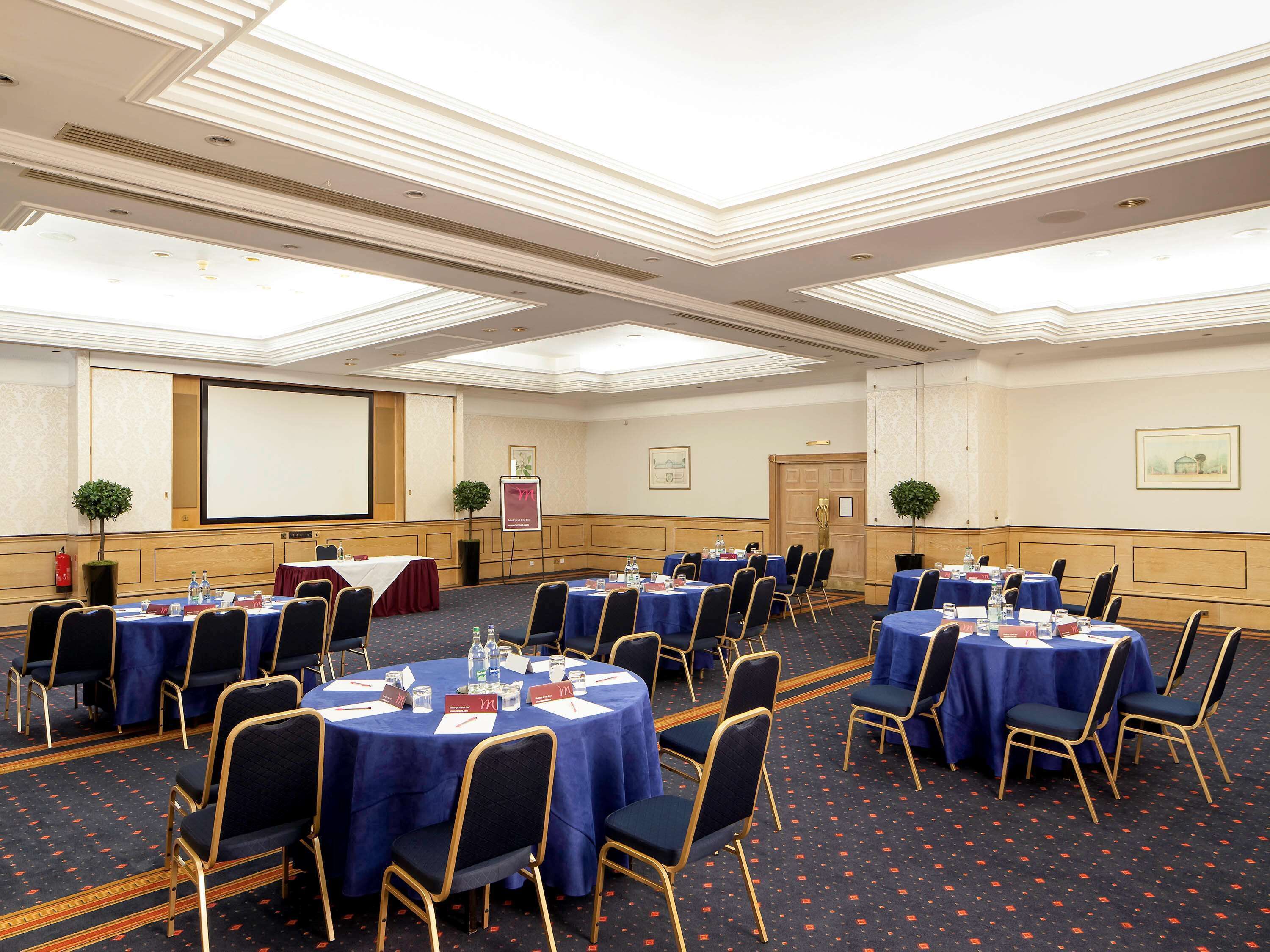 Une salle de conférence équipée de tables rondes, de chaises, d’un écran de projection et d’un tableau de présentation.
