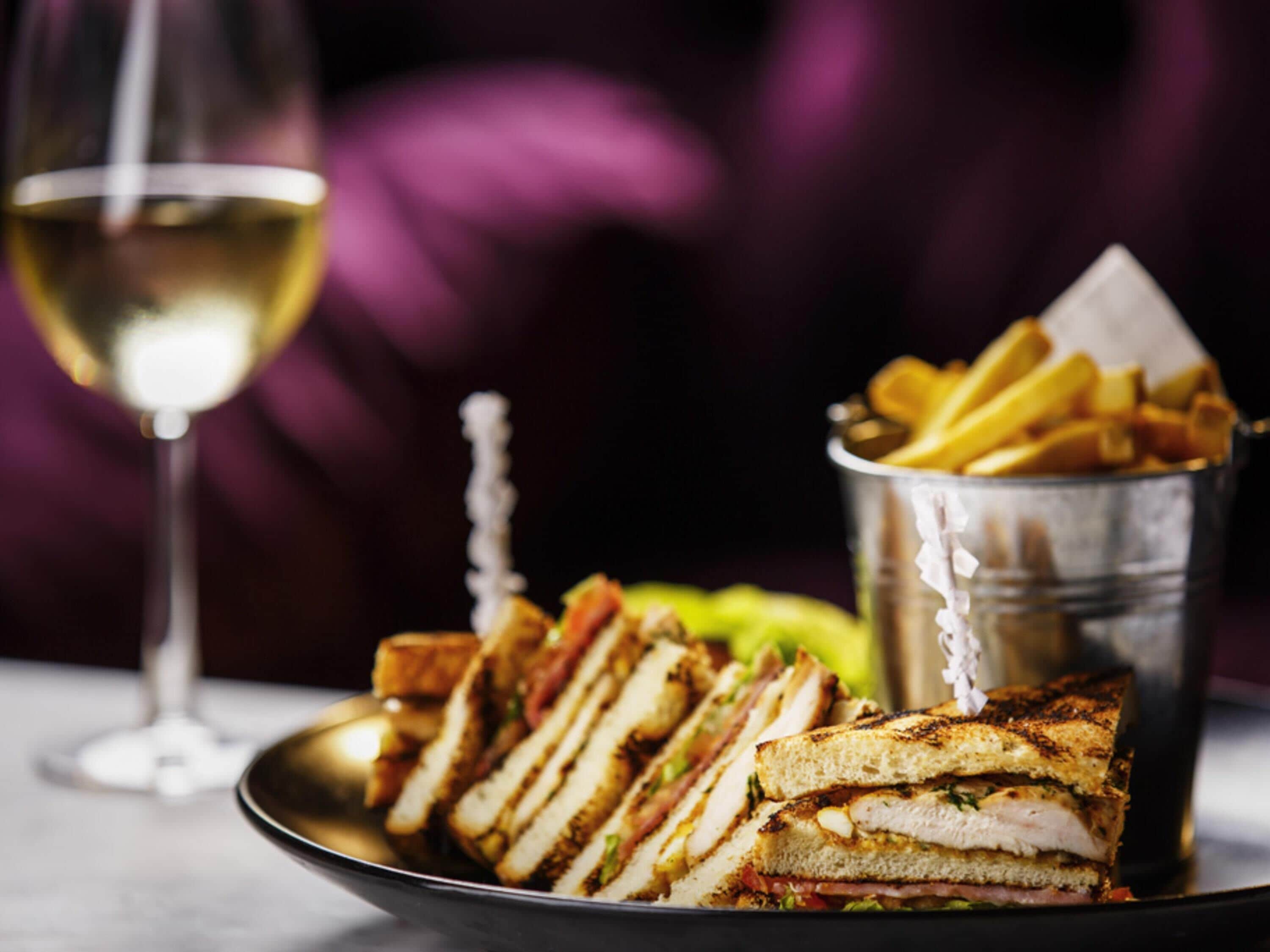 Une assiette de sandwichs grillés et un seau de frites, avec un verre de vin blanc en arrière-plan.