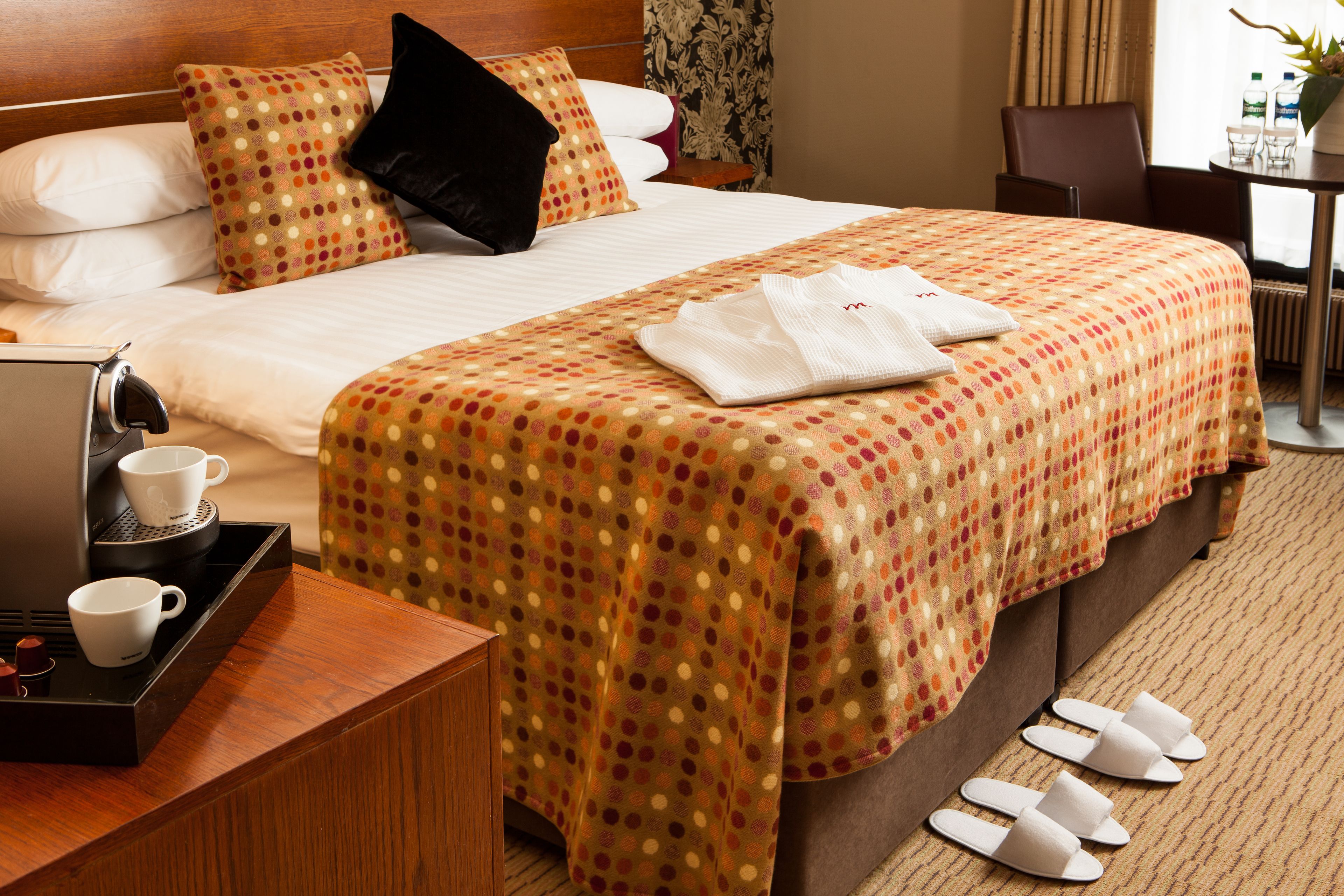 A hotel room with a neatly made bed, a coffee maker, a tray with cups, slippers, and a small table with a plant.