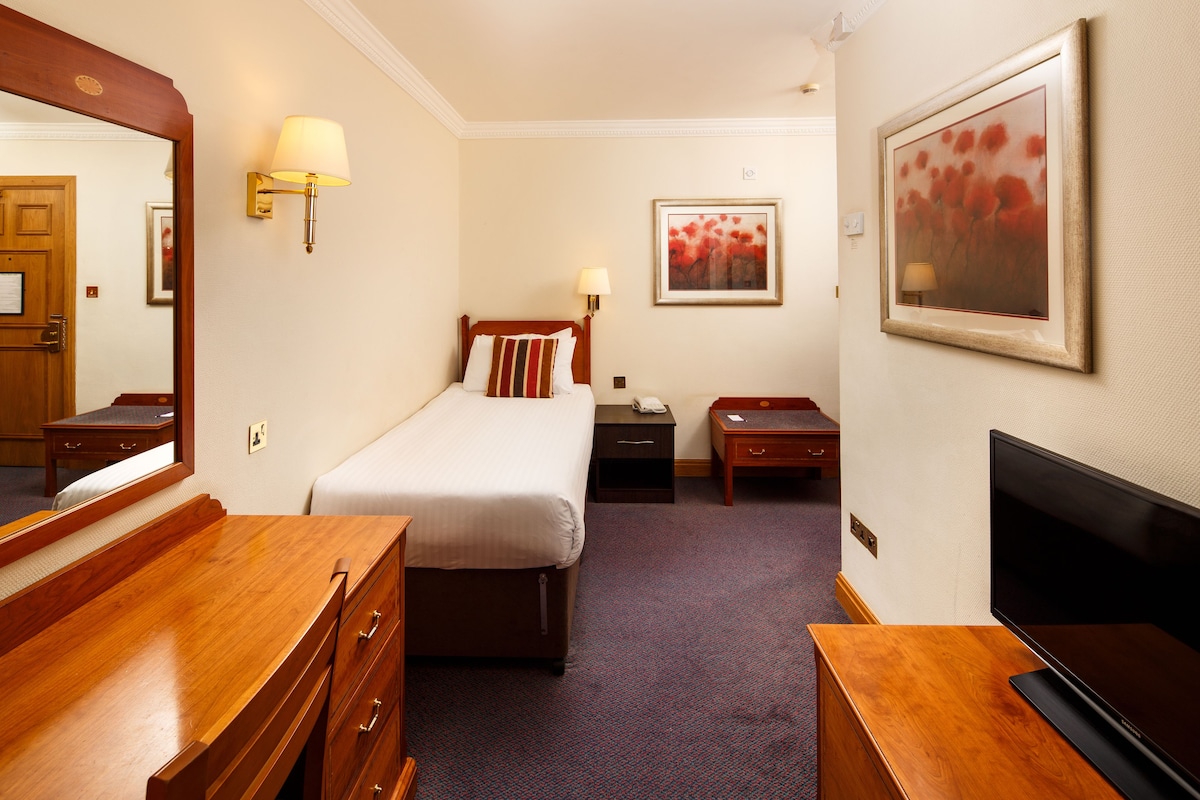 A hotel room with a bed, bedside tables, a dresser, a TV, and a painting on the wall.