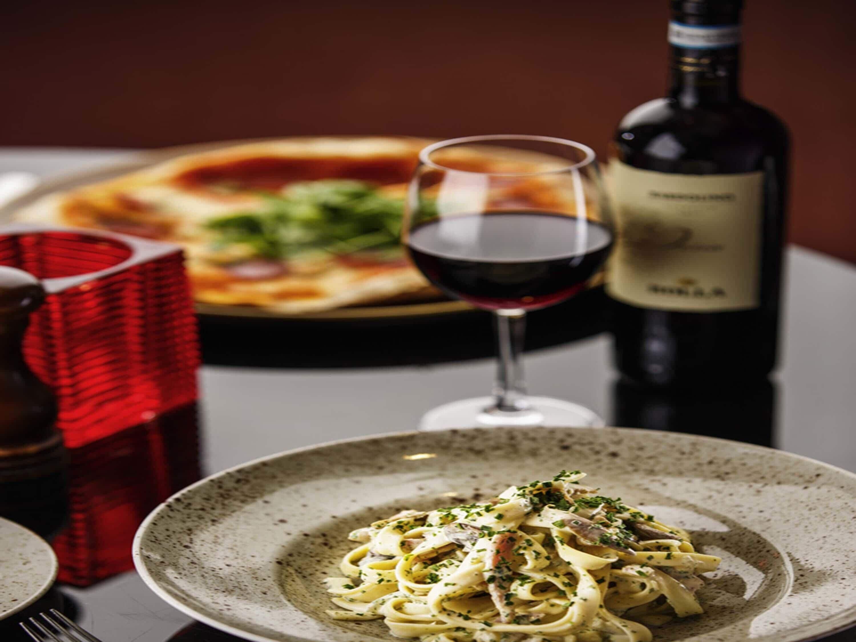 Una mesa de comedor con un plato de pasta, una copa de vino tinto y una botella de vino.