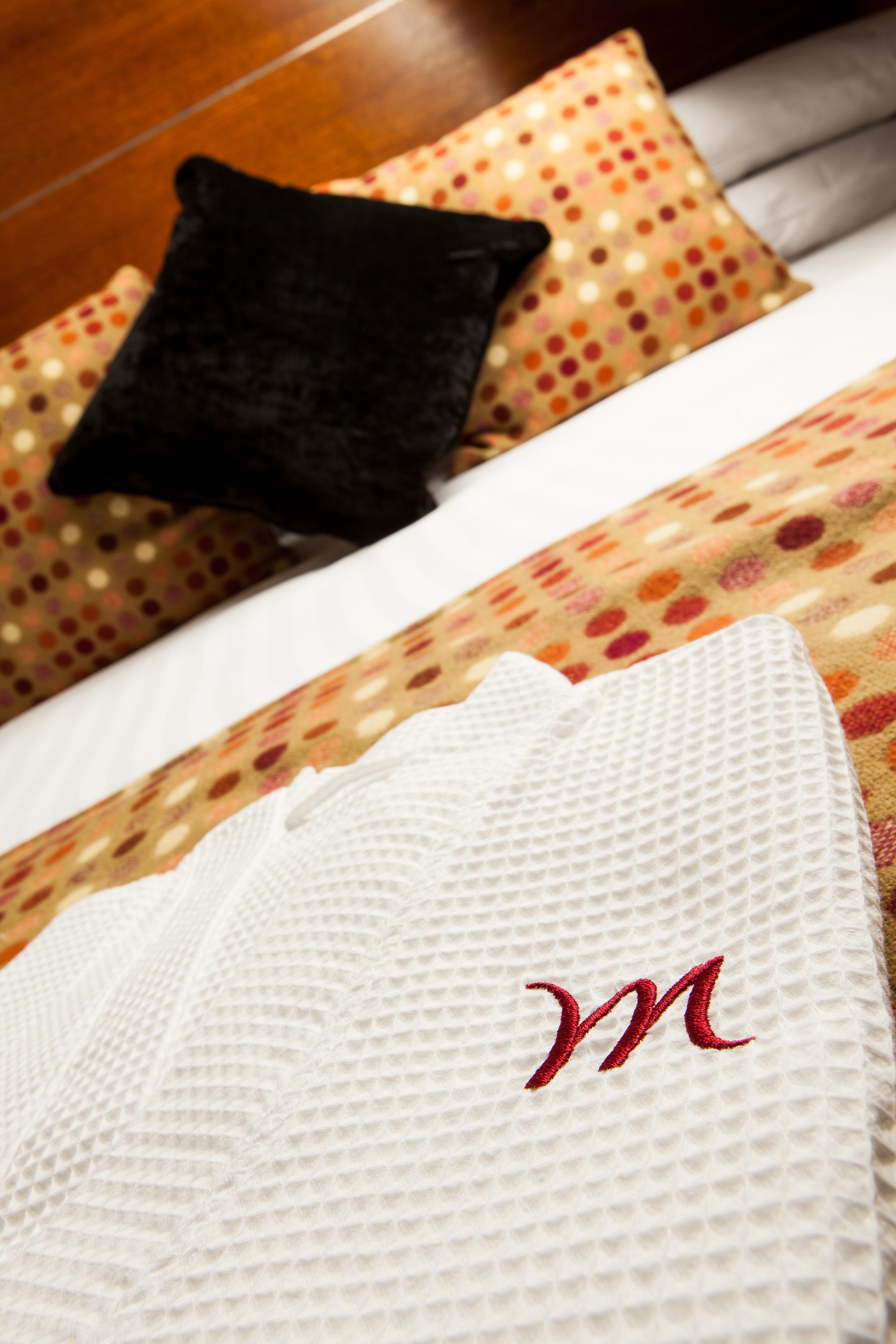 A neatly made bed with a white pillow featuring a red 'M' embroidery, a black pillow, and a polka-dotted pillow.