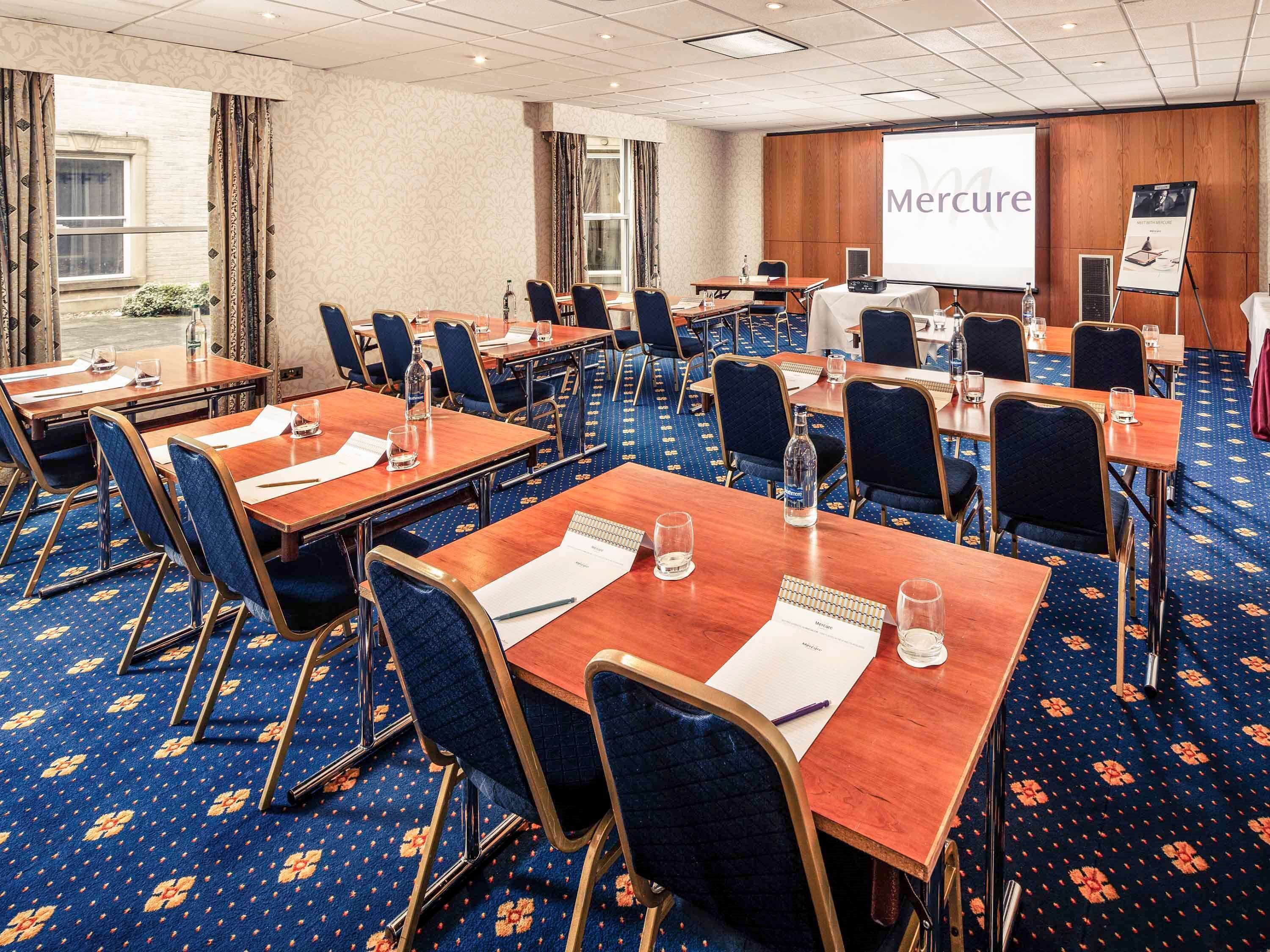A conference room with multiple tables and chairs, a projector screen displaying 'Mercure', and a presentation board.