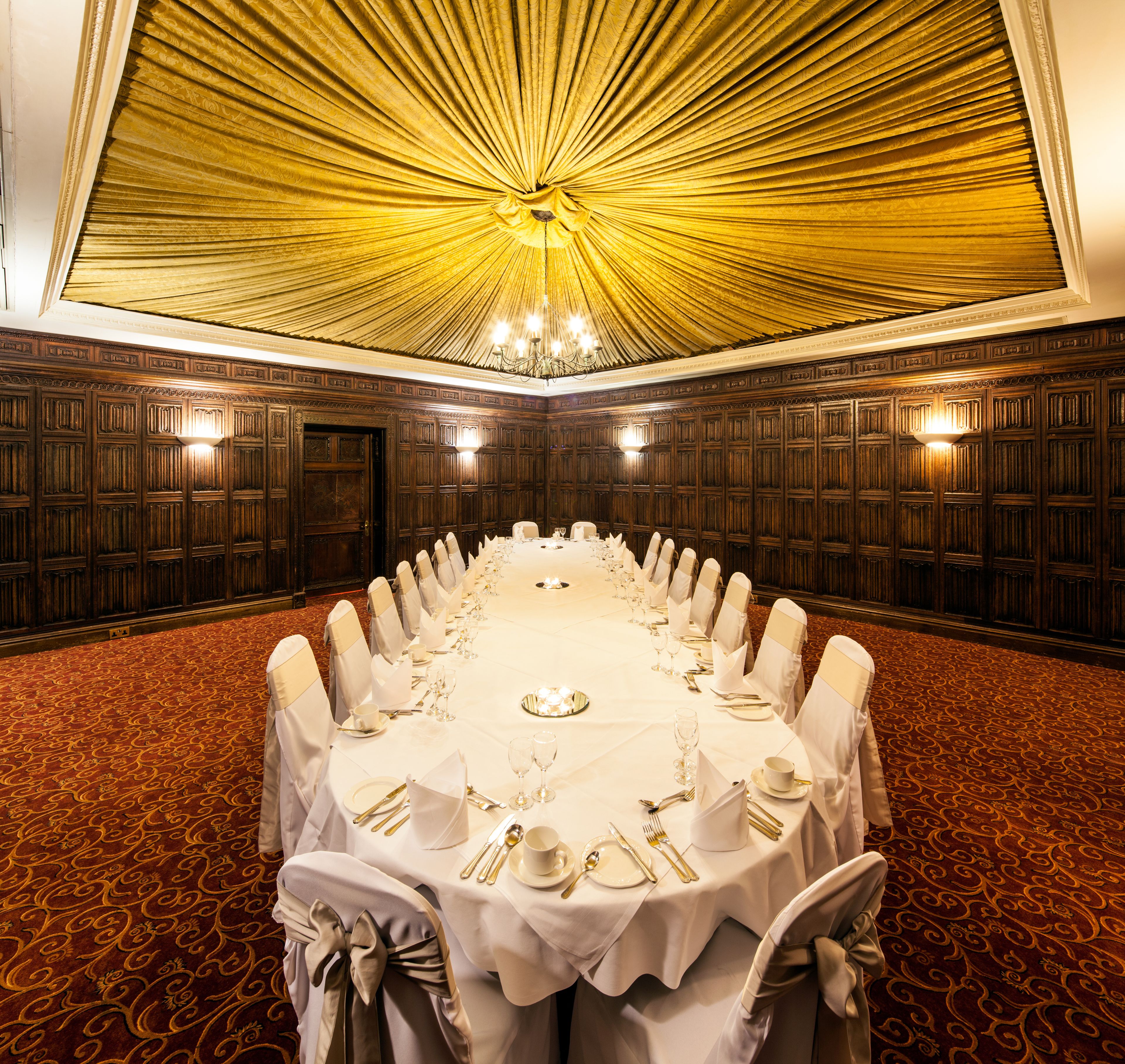 A conference room with a long table set for a formal event, surrounded by wooden paneling and a patterned carpet.