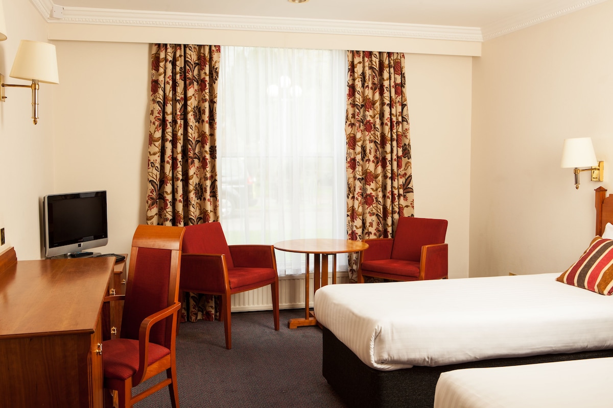 A hotel room with a bed, two red chairs, a small table, a TV, and curtains.