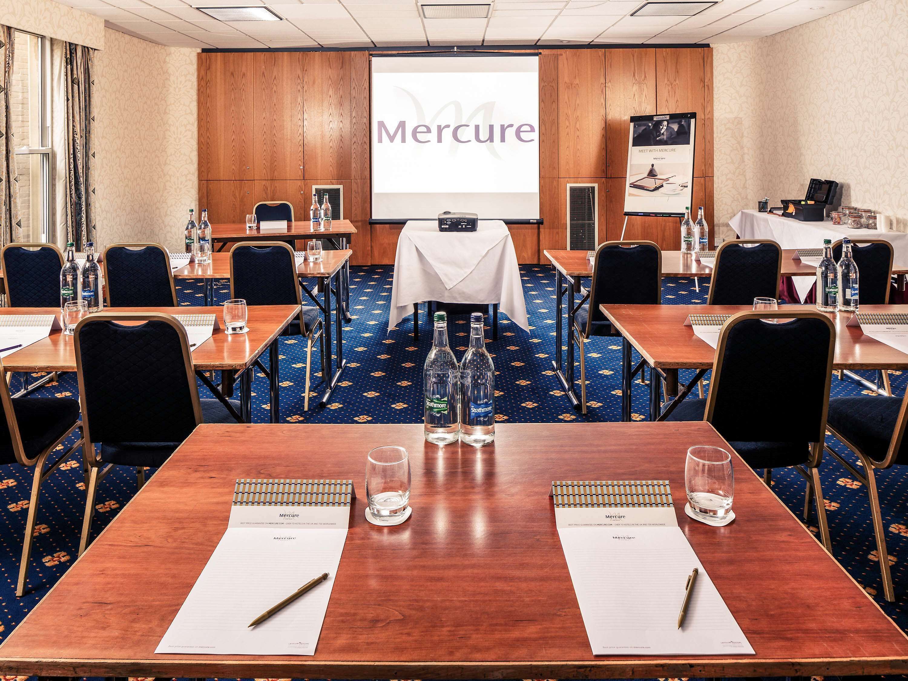 Sala de conferência com uma grande tela exibindo a marca 'Mercure', várias mesas com cadeiras, garrafas de água e cadernos.
