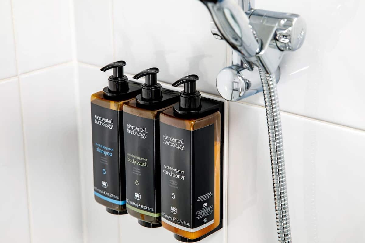 Three bottles of Elemental Herbology shampoo, body wash, and conditioner mounted on a wall near a shower.