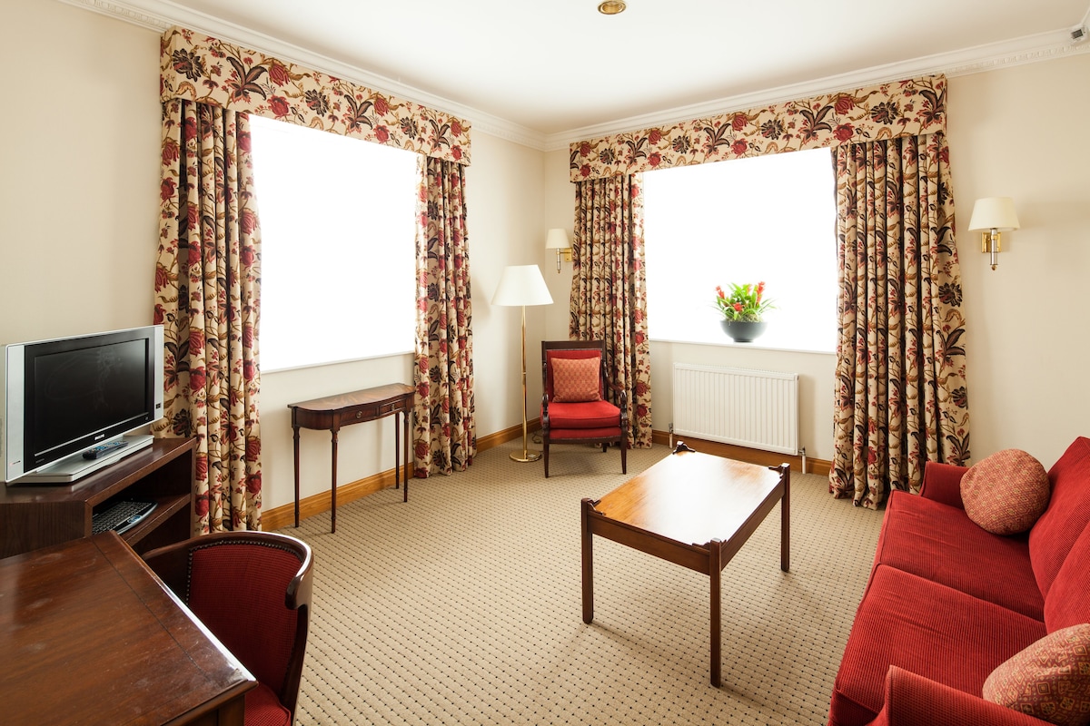 A hotel room with a TV, a red sofa, a wooden coffee table, a red chair, a floor lamp, and floral curtains.
