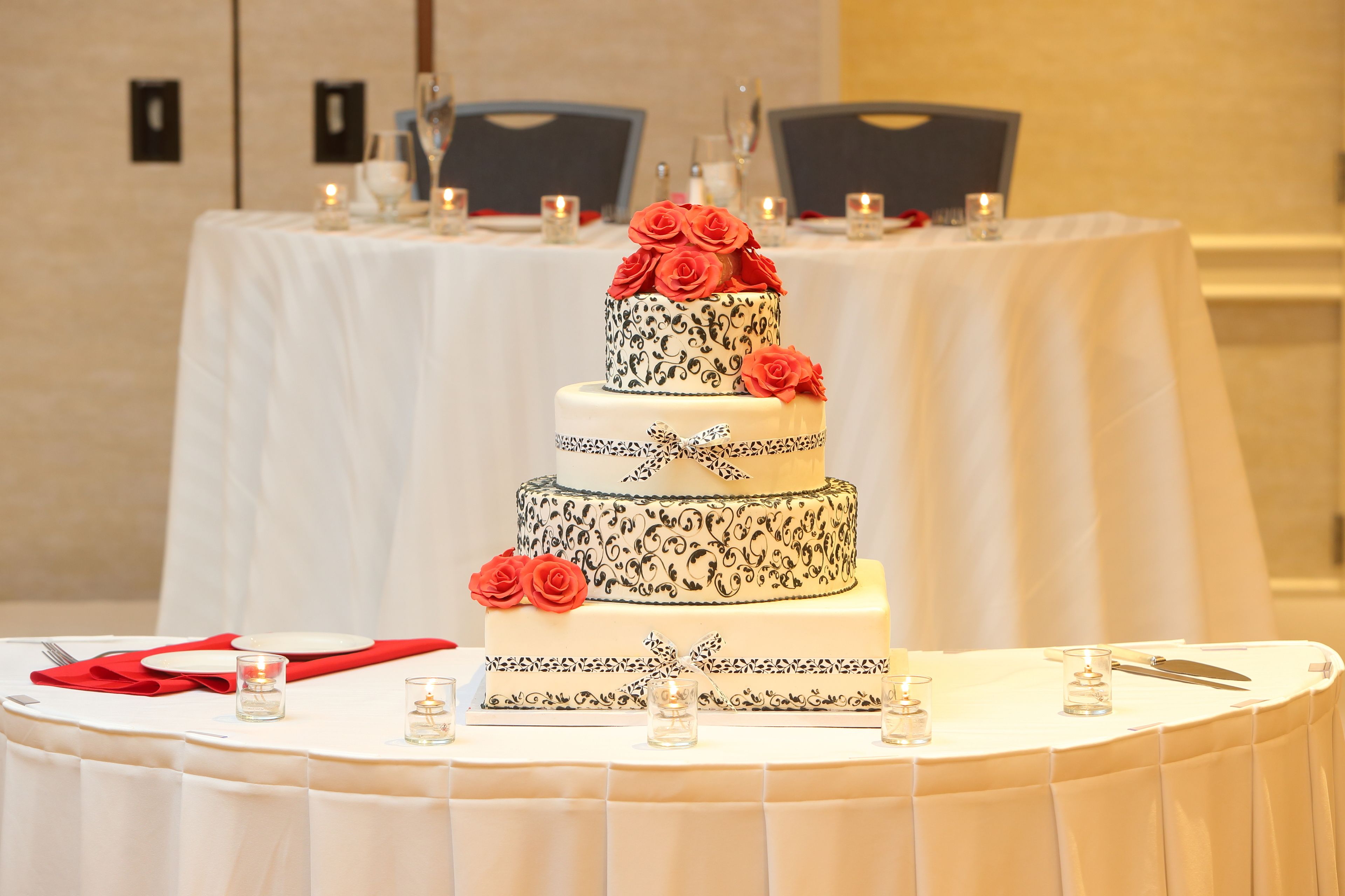 A three-tiered wedding cake with black and white patterns and red roses on top, placed on a round table with white tablecloths.