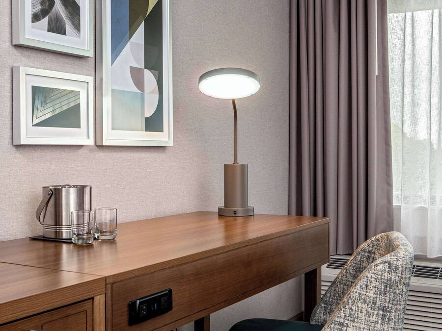 Habitación de hotel con un escritorio de madera, una lámpara moderna, un hervidor y dos vasos. Hay pinturas abstractas en la pared y una ventana con cortinas.