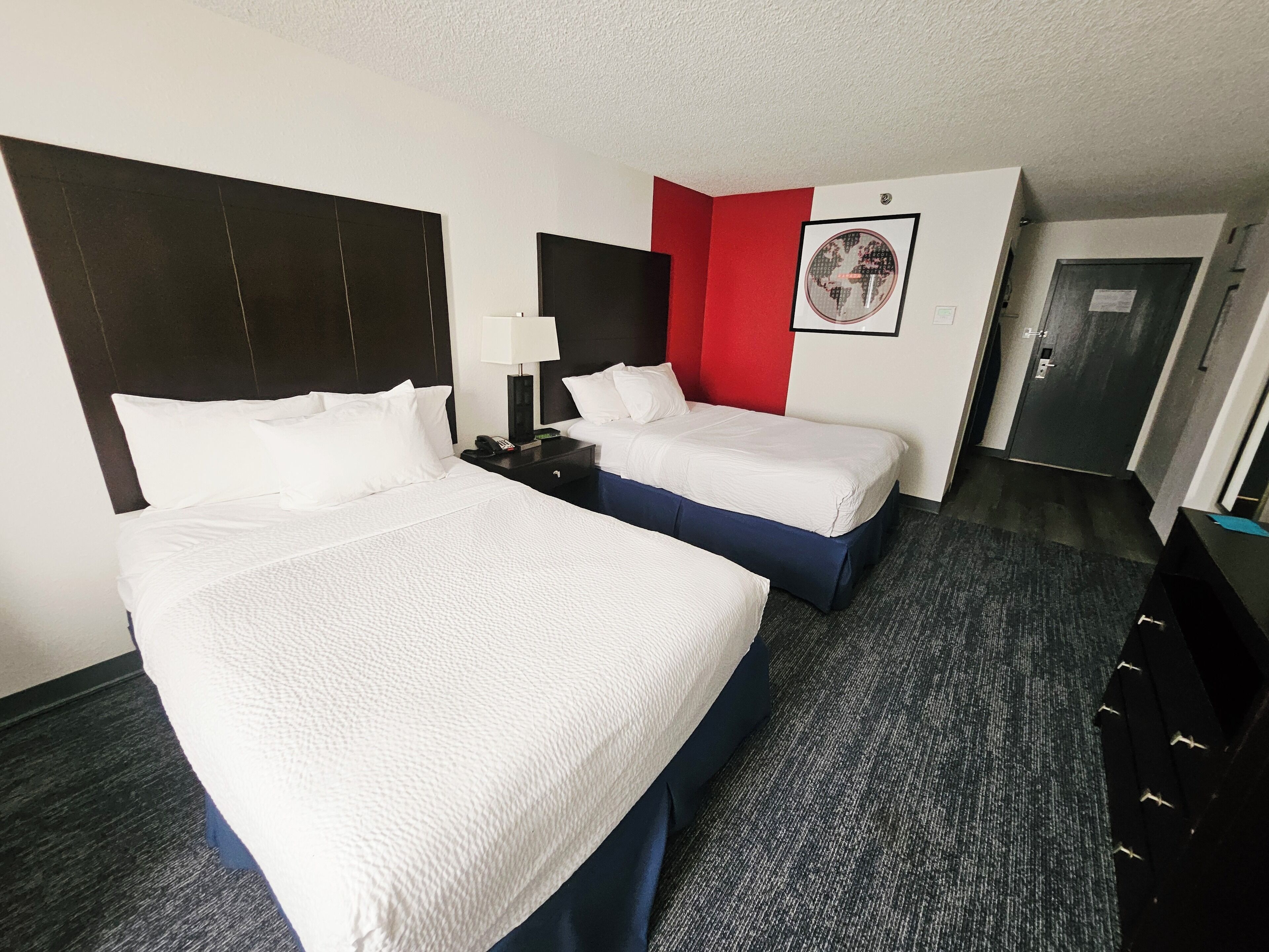 A hotel room with two beds, a red accent wall, a framed picture, and a door.