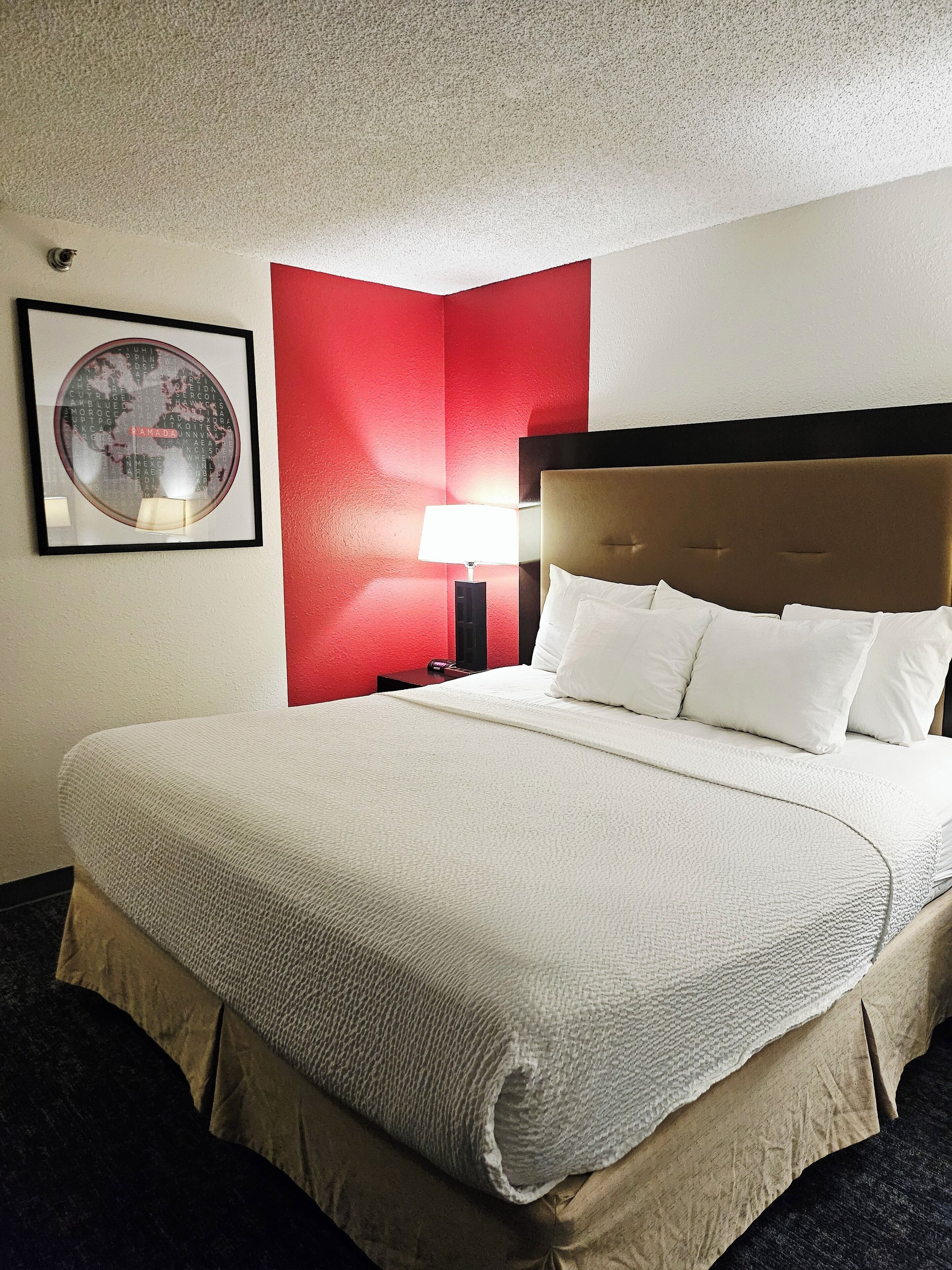 A hotel room with a large bed, a red accent wall, a framed world map, and a nightstand with a lamp.