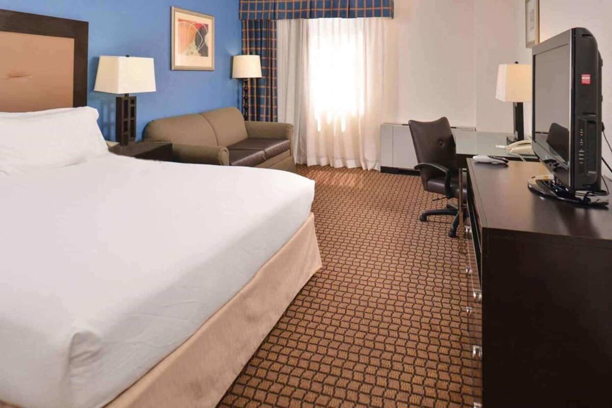 A hotel room with a large bed, a desk with a computer, a chair, a sofa, and a window with curtains.
