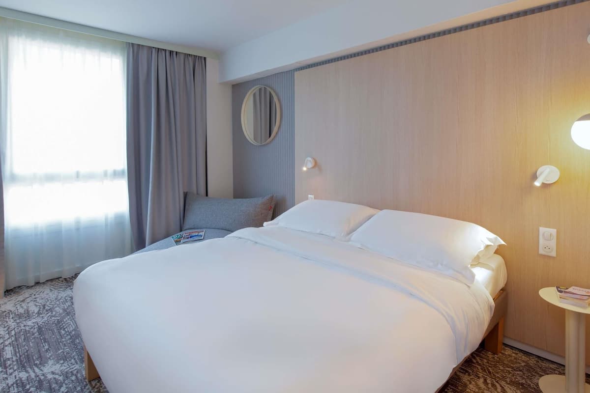 A hotel room with a large bed, a round mirror, a small table with a magazine, and a window with curtains.