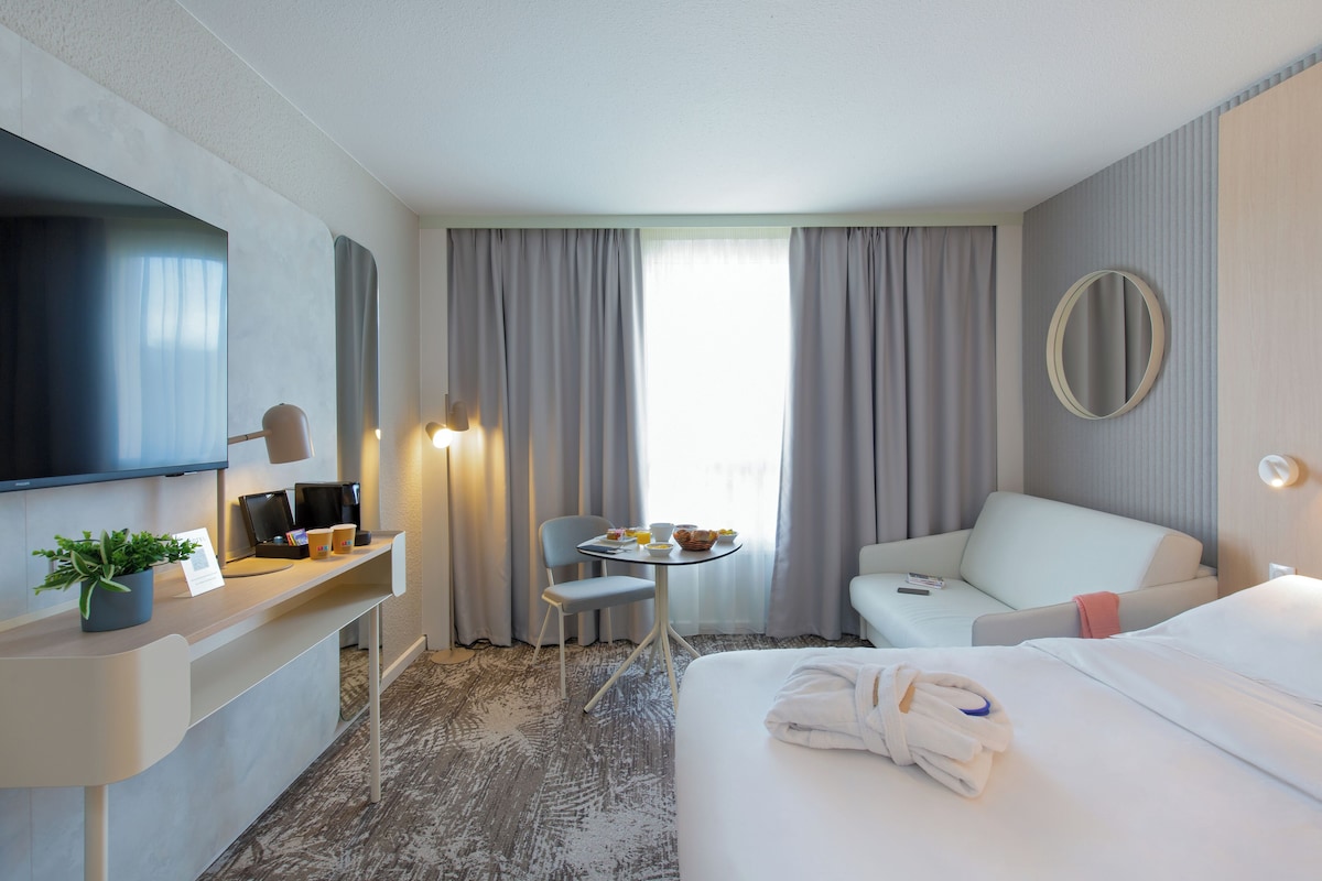Une chambre d’hôtel comprenant un lit, un bureau avec un ordinateur, une chaise, une petite table avec des articles pour le petit-déjeuner, une télévision et une fenêtre avec des rideaux.