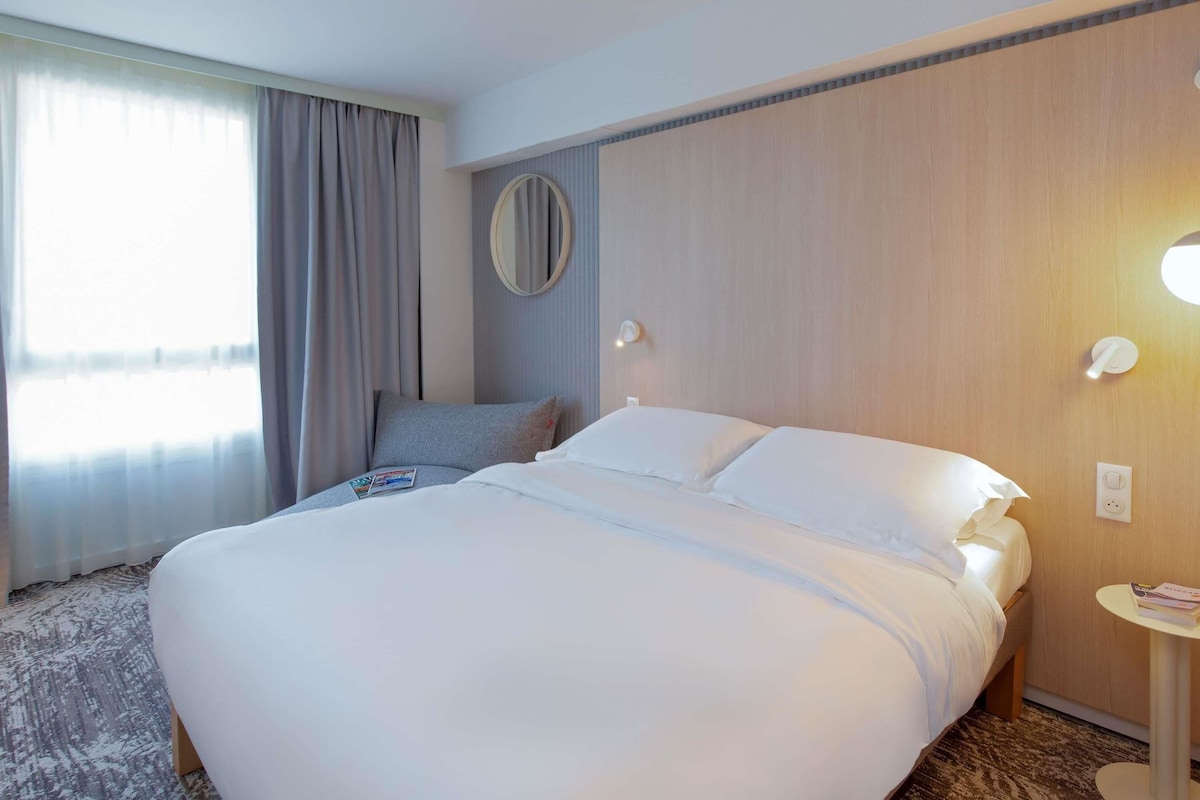 Une chambre d’hôtel avec un grand lit, un miroir rond, une petite table avec un magazine et une fenêtre avec des rideaux.