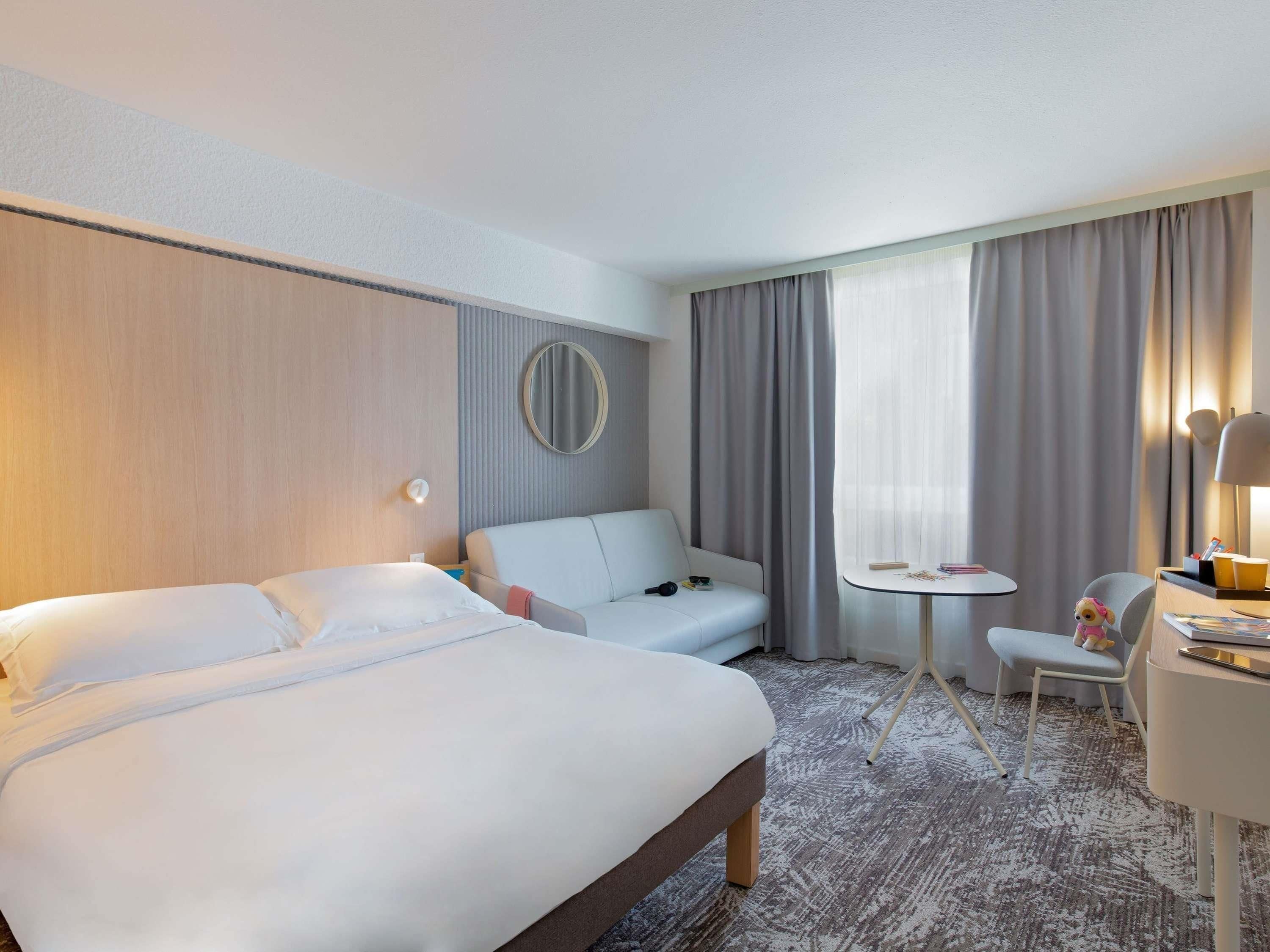 Habitación de hotel con cama, sofá, una mesita, una silla, una lámpara y una ventana con cortinas.
