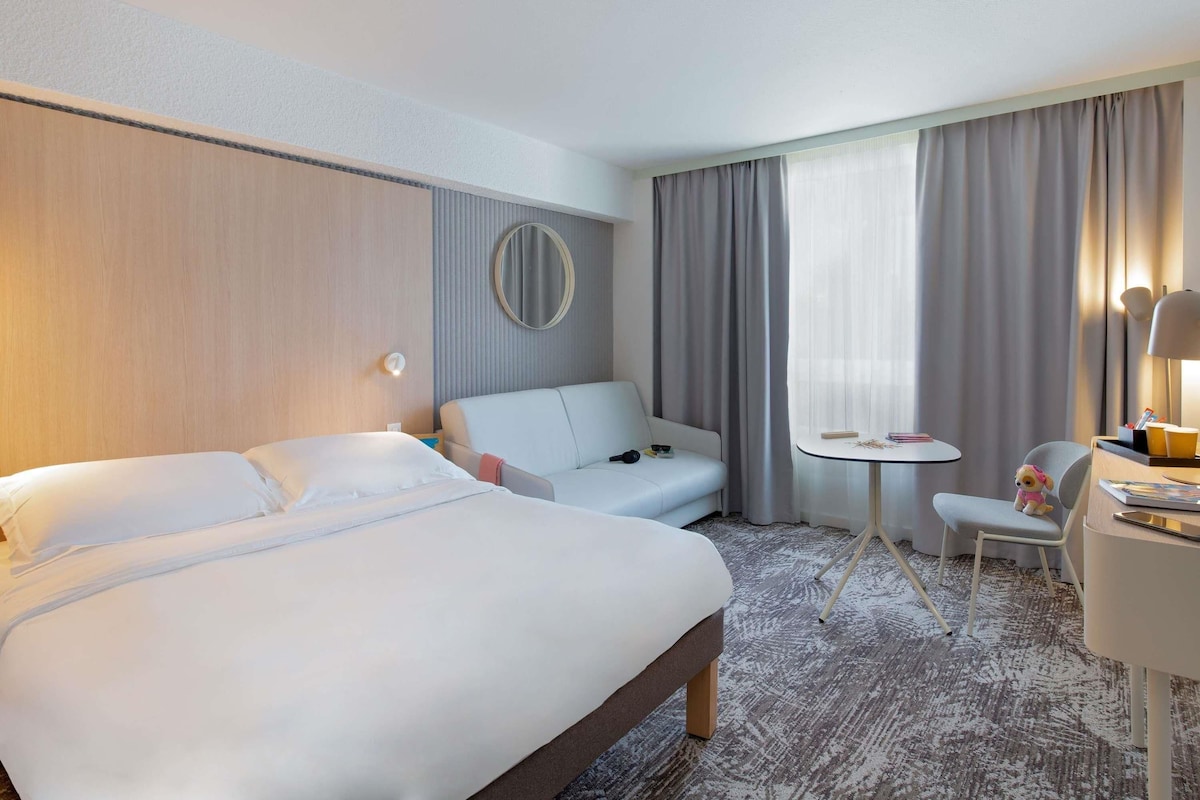 Une chambre d’hôtel avec un lit, un canapé, une petite table, une chaise, une lampe et une fenêtre avec des rideaux.
