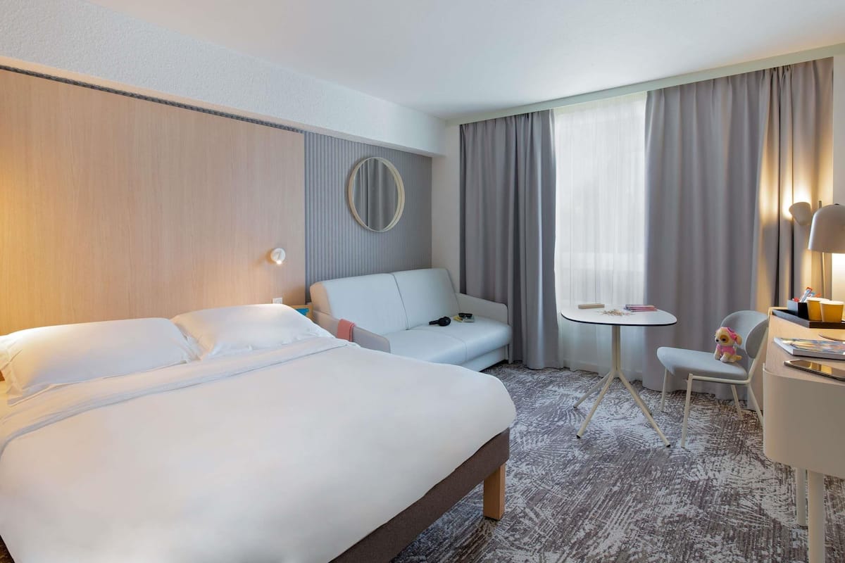 A hotel room with a bed, a sofa, a small table, a chair, a lamp, and a window with curtains.