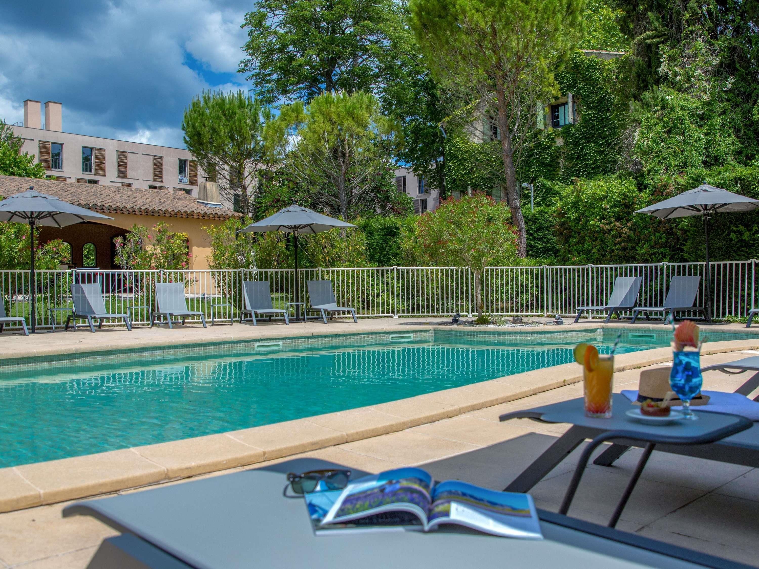 Un espace piscine doté de chaises longues et de parasols, entouré d’arbres et d’un bâtiment.