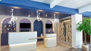 A reception area with a modern design, featuring a reception desk, a flat-screen TV, and a decorative plant. - Hotel Novotel Aix en Provence Pont de l'Arc Fenouillères (Aix-en-Provence)