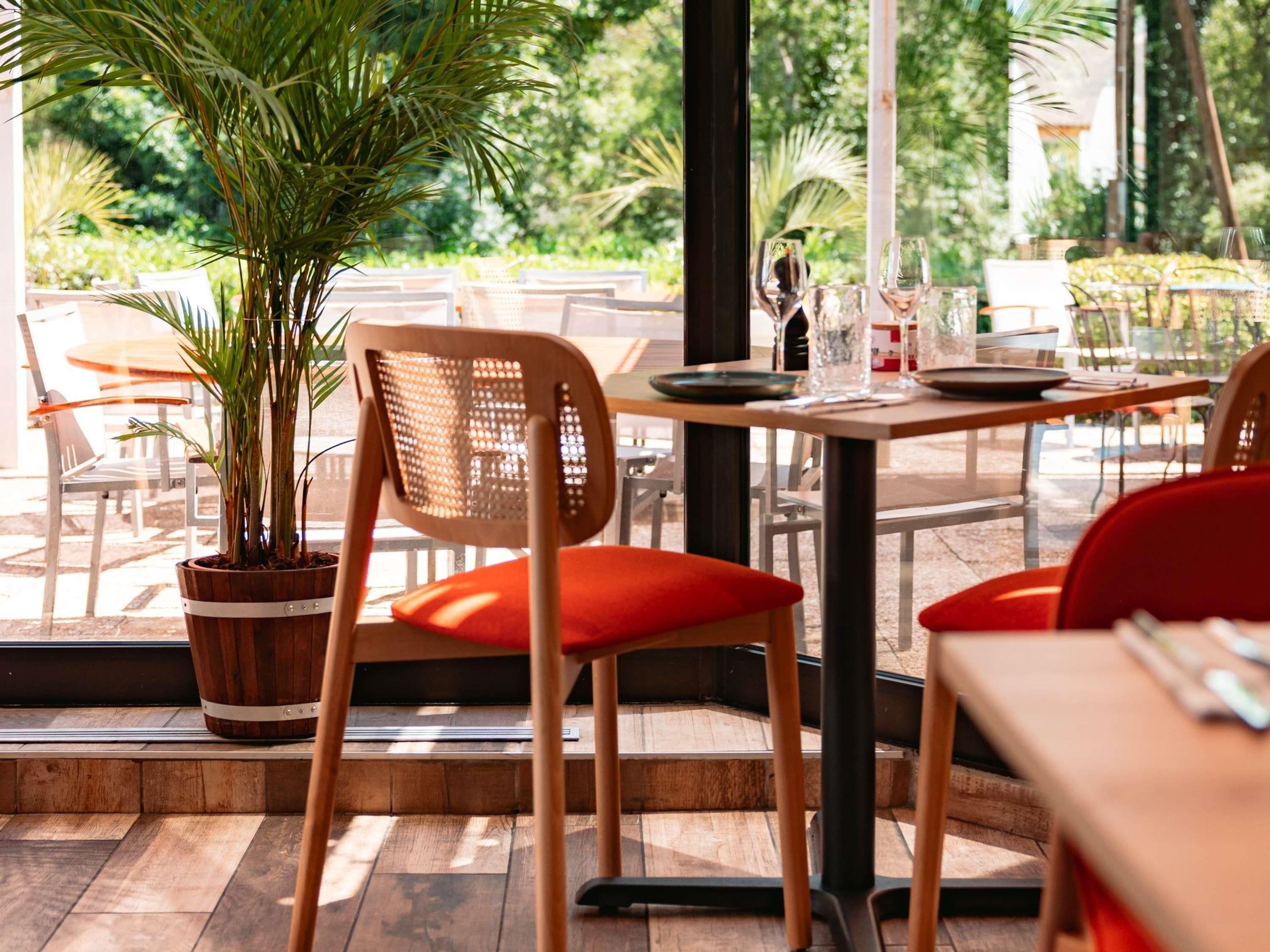 An outdoor dining area with wooden tables and chairs, a potted plant, and a view of greenery.