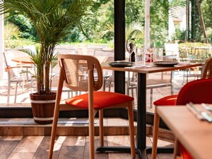 An outdoor dining area with wooden tables and chairs, a potted plant, and a view of greenery.