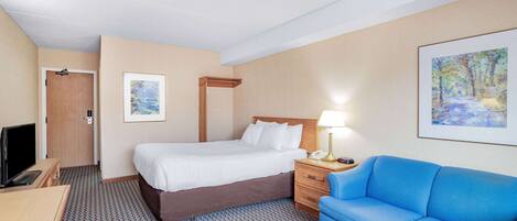 A hotel room with a bed, a blue sofa, a desk with a TV, and a painting on the wall.