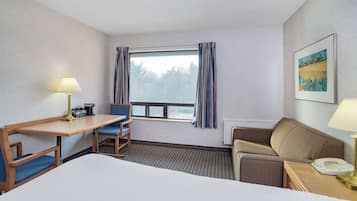 Quarto de hotel com cama, escrivaninha com luminária, poltrona, sofá, janela com cortinas e um quadro na parede.