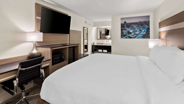 A hotel room with a large bed, a desk with a chair, a flat-screen TV, a cityscape painting, and a mirror. - Clarion Pointe Kennesaw Town Center (Kennesaw)