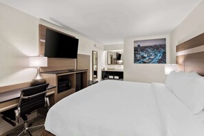 A hotel room with a large bed, a desk with a chair, a flat-screen TV, a cityscape painting, and a mirror.