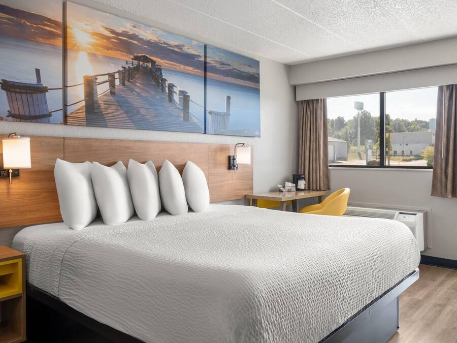 Habitación de hotel con una cama grande, cabecera de madera, un cuadro de un muelle, un escritorio con una silla y una ventana con cortinas.