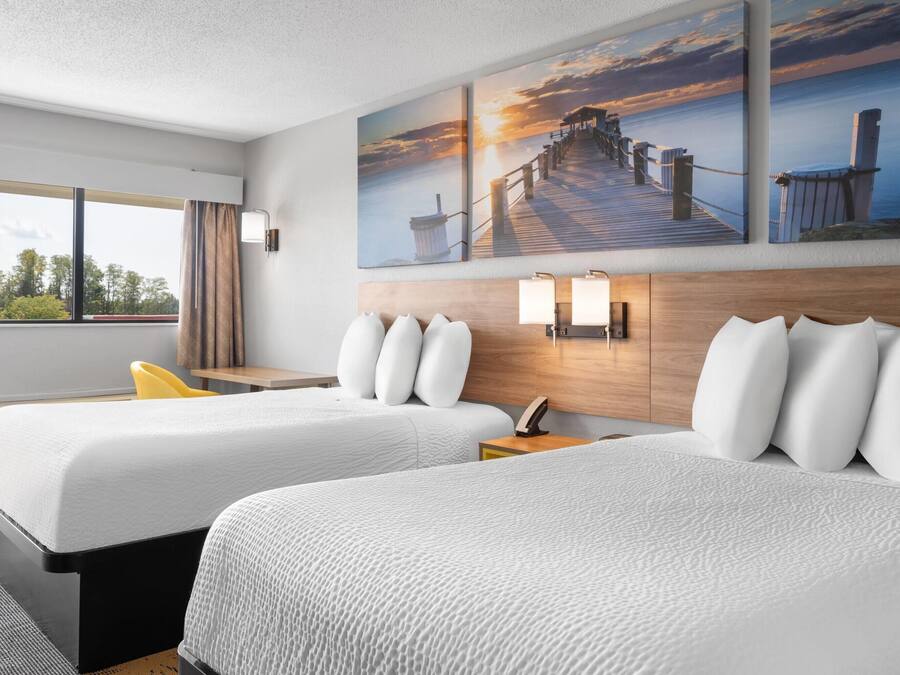 A hotel room with two beds, a wooden headboard, a nightstand with a lamp, and a window with curtains.