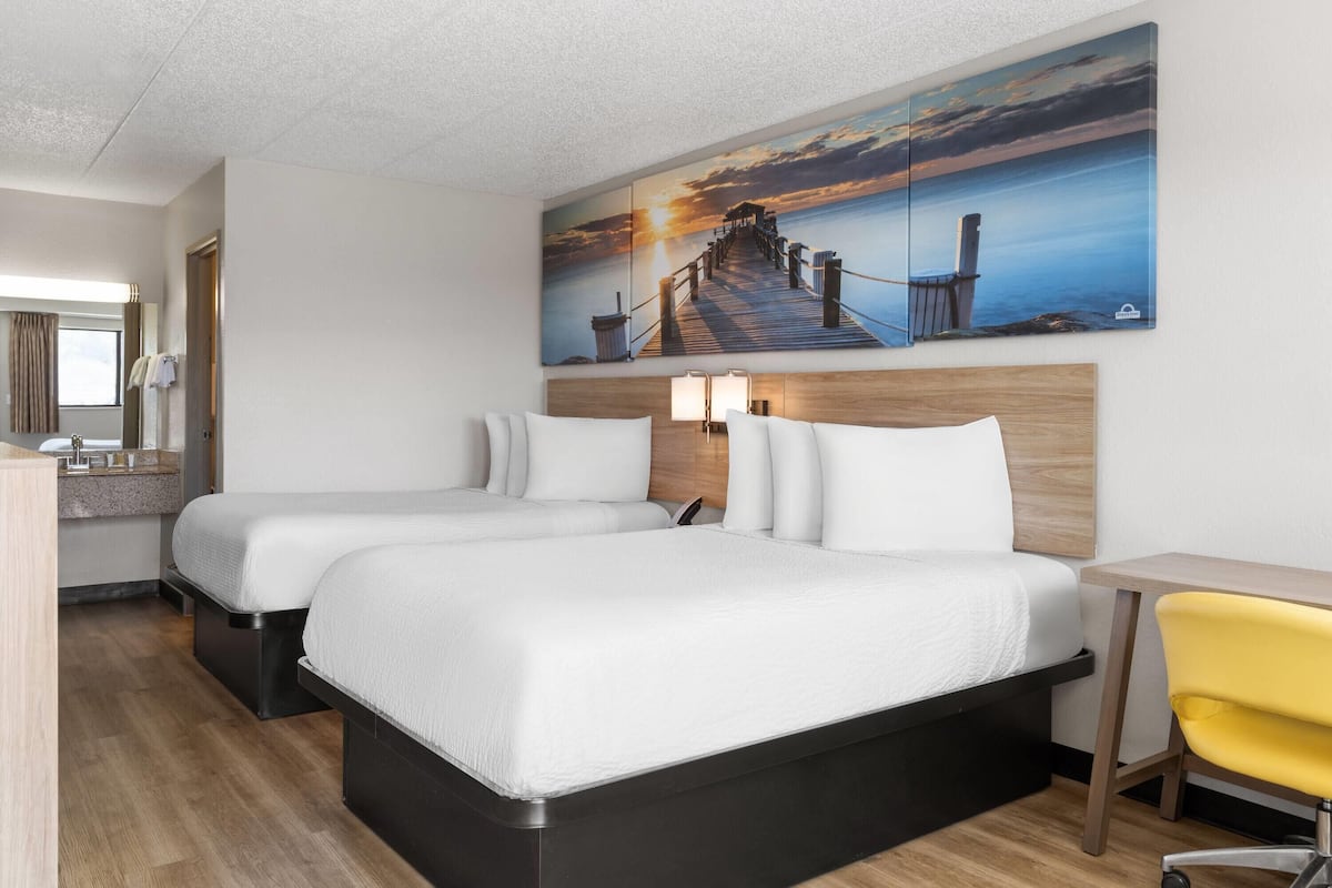A hotel room with two beds, a desk, a chair, and a large painting of a sunset over a pier.