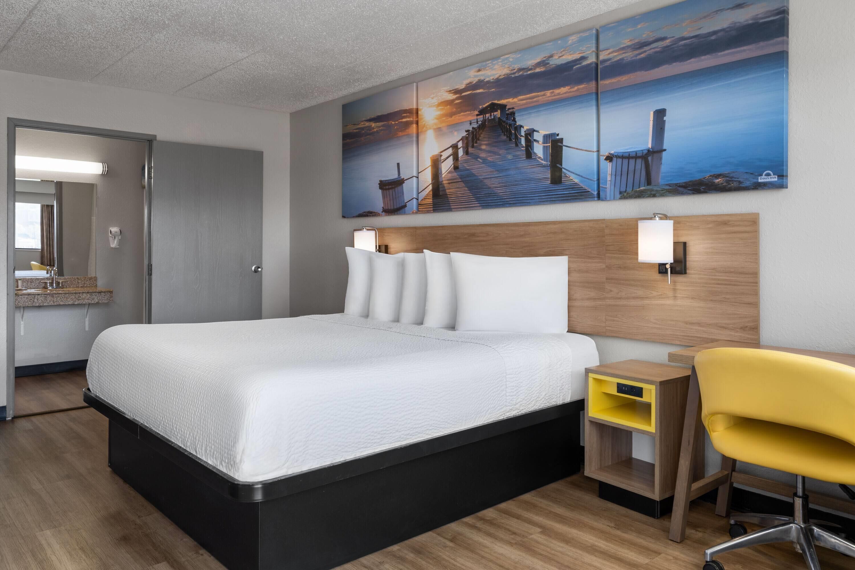 Quarto de hotel com uma cama grande, cabeceira de madeira, mesa de cabeceira, uma cadeira amarela e um quadro de um cais na parede.