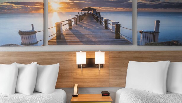 A hotel room with a wooden headboard, two white beds, a yellow nightstand, a wall-mounted lamp, and a painting of a pier at sunset. - Days Inn by Wyndham Cloverdale Greencastle (Cloverdale)