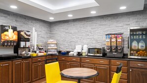 A modern kitchen area with a brick wall, wooden cabinets, and a coffee machine. There are two yellow chairs around a small wooden table. - Days Inn by Wyndham Cloverdale Greencastle (Cloverdale)