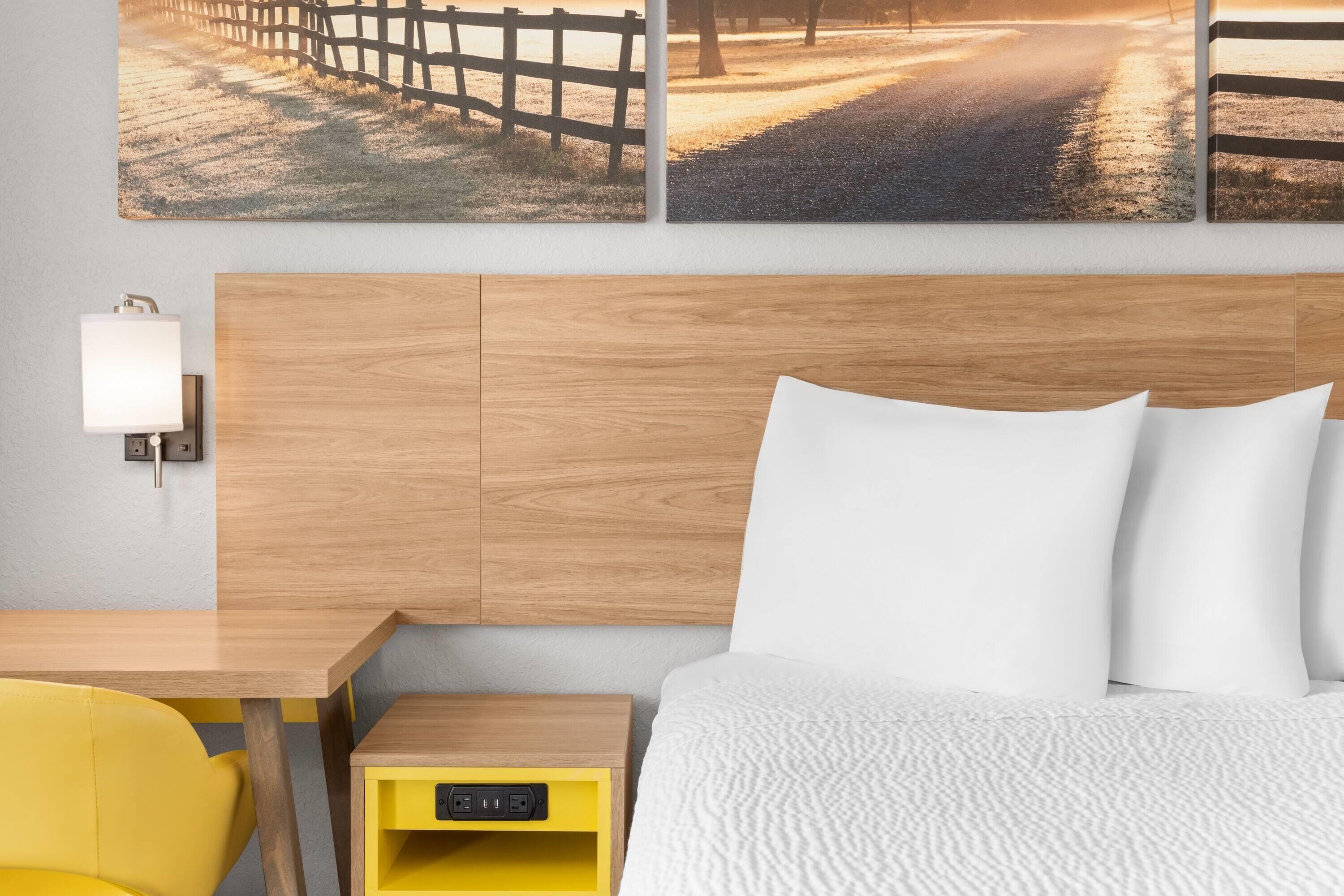 Habitación de hotel con cabecera de madera, cama con ropa blanca, mesita de noche amarilla con lámpara y una obra de arte en la pared que representa una escena rural.