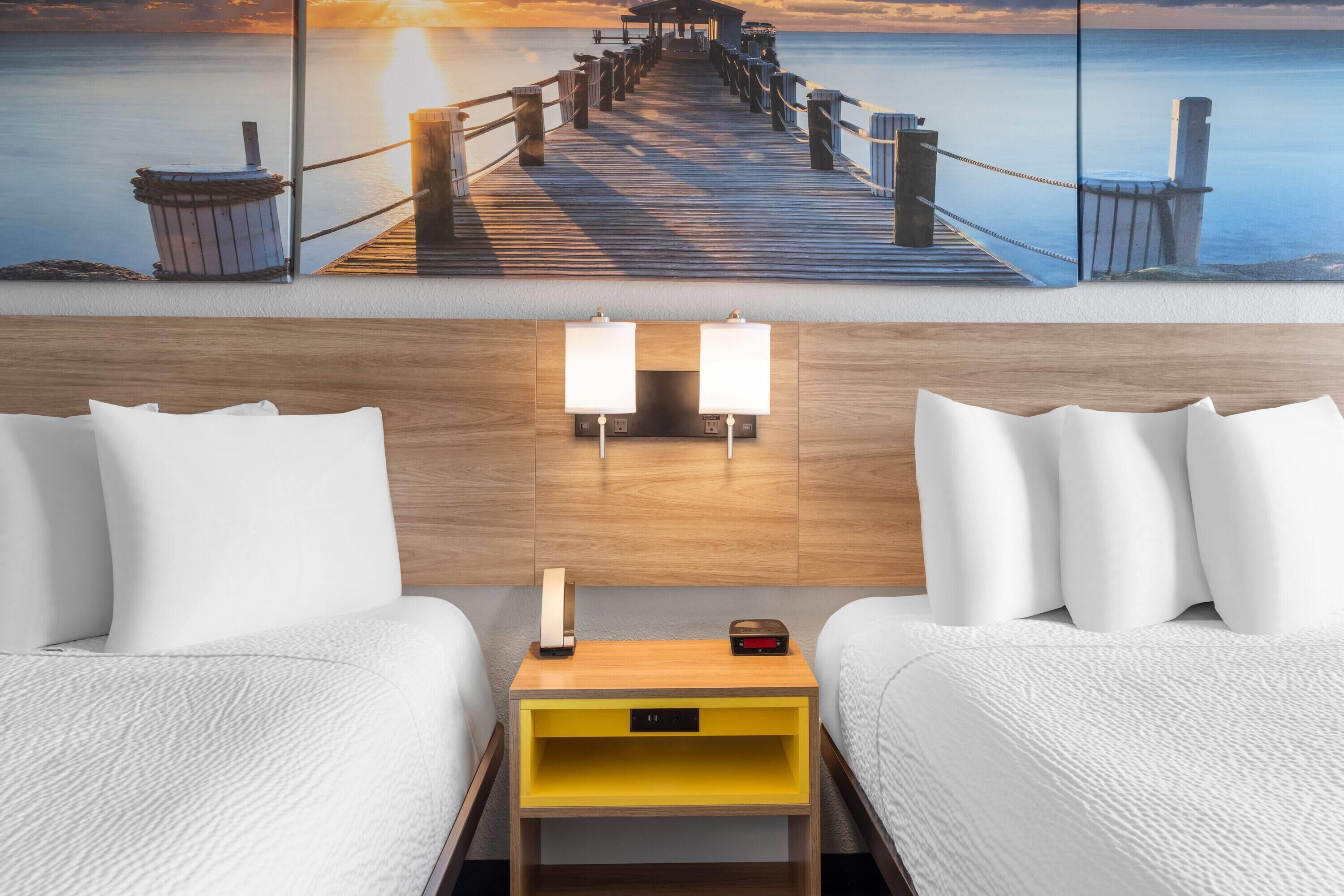 A hotel room with a wooden headboard, two white beds, a yellow nightstand with a lamp, and a painting of a pier at sunset.