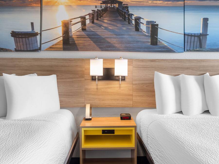 A hotel room with a wooden headboard, two white beds, a yellow nightstand with a lamp, and a painting of a pier at sunset.
