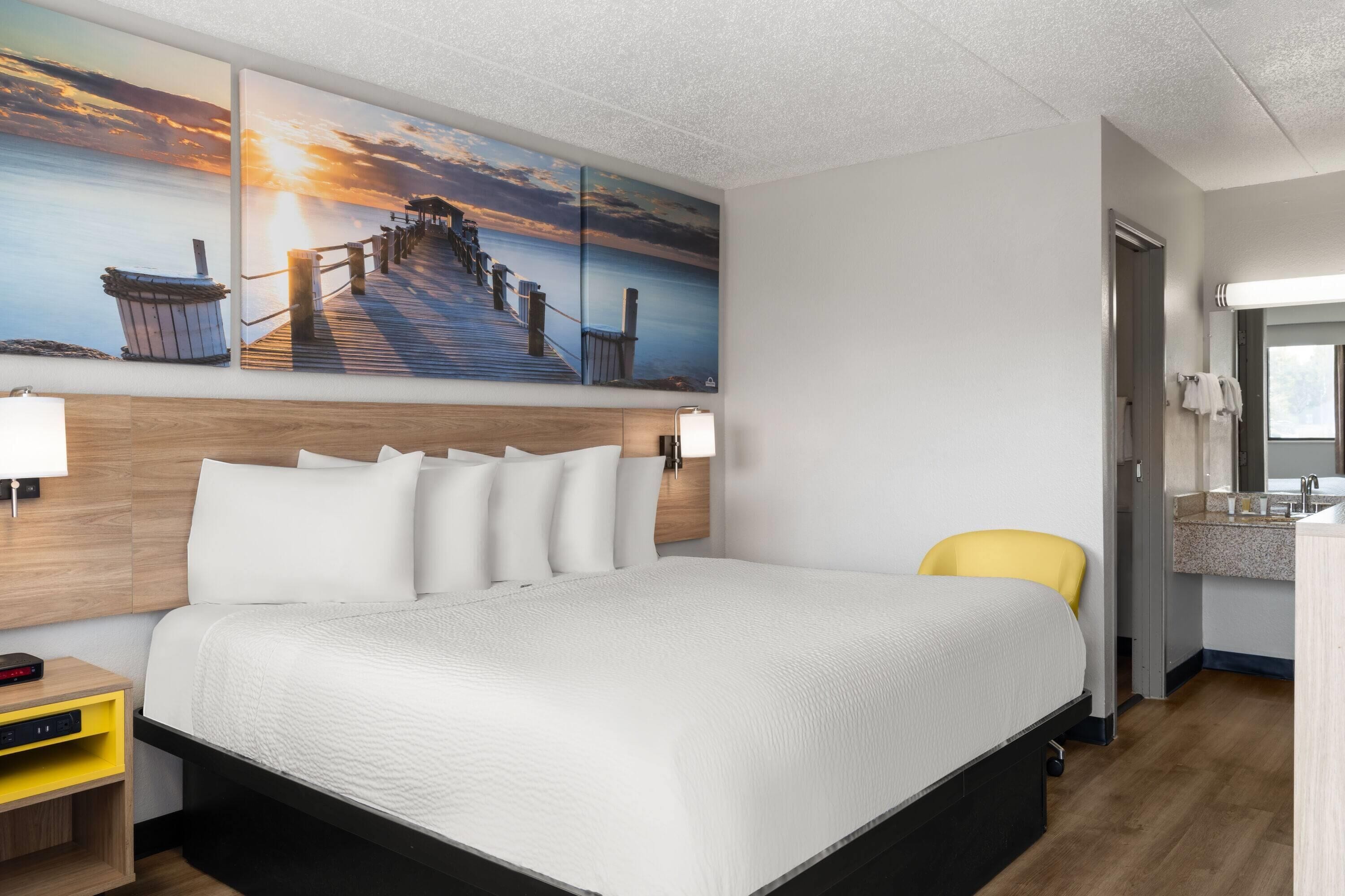 A hotel room with a large bed, a wooden headboard, a painting of a pier, a yellow chair, and a bathroom with a sink and mirror.