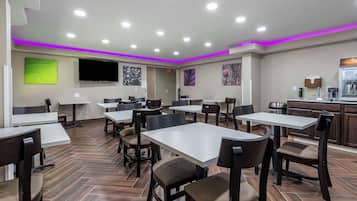 Un restaurante con varias mesas y sillas, un televisor de pantalla plana montado en la pared y una zona de cocina visible al fondo.