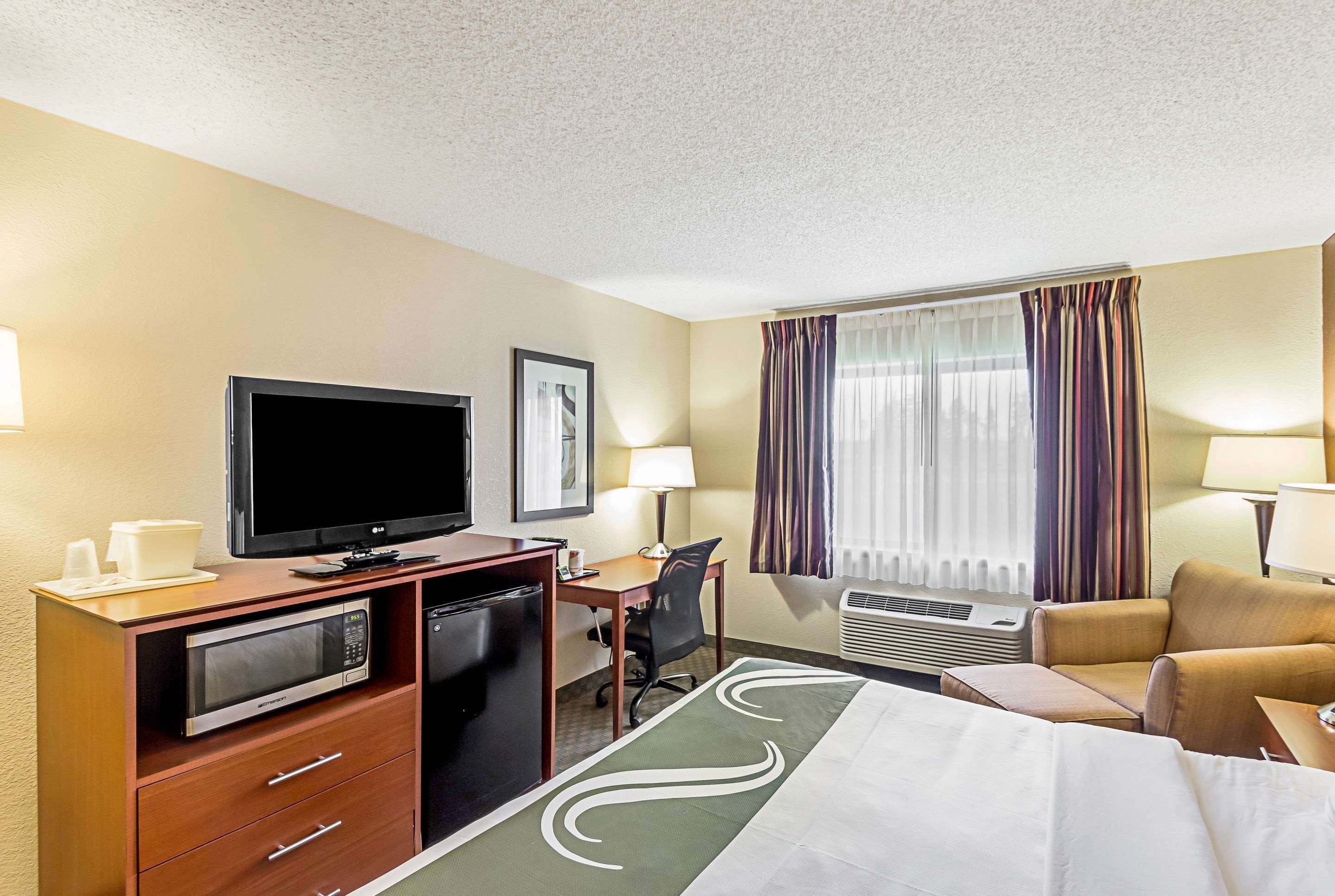 A hotel room with a bed, a desk with a computer, a television, a microwave, a mini-fridge, and a window with curtains.