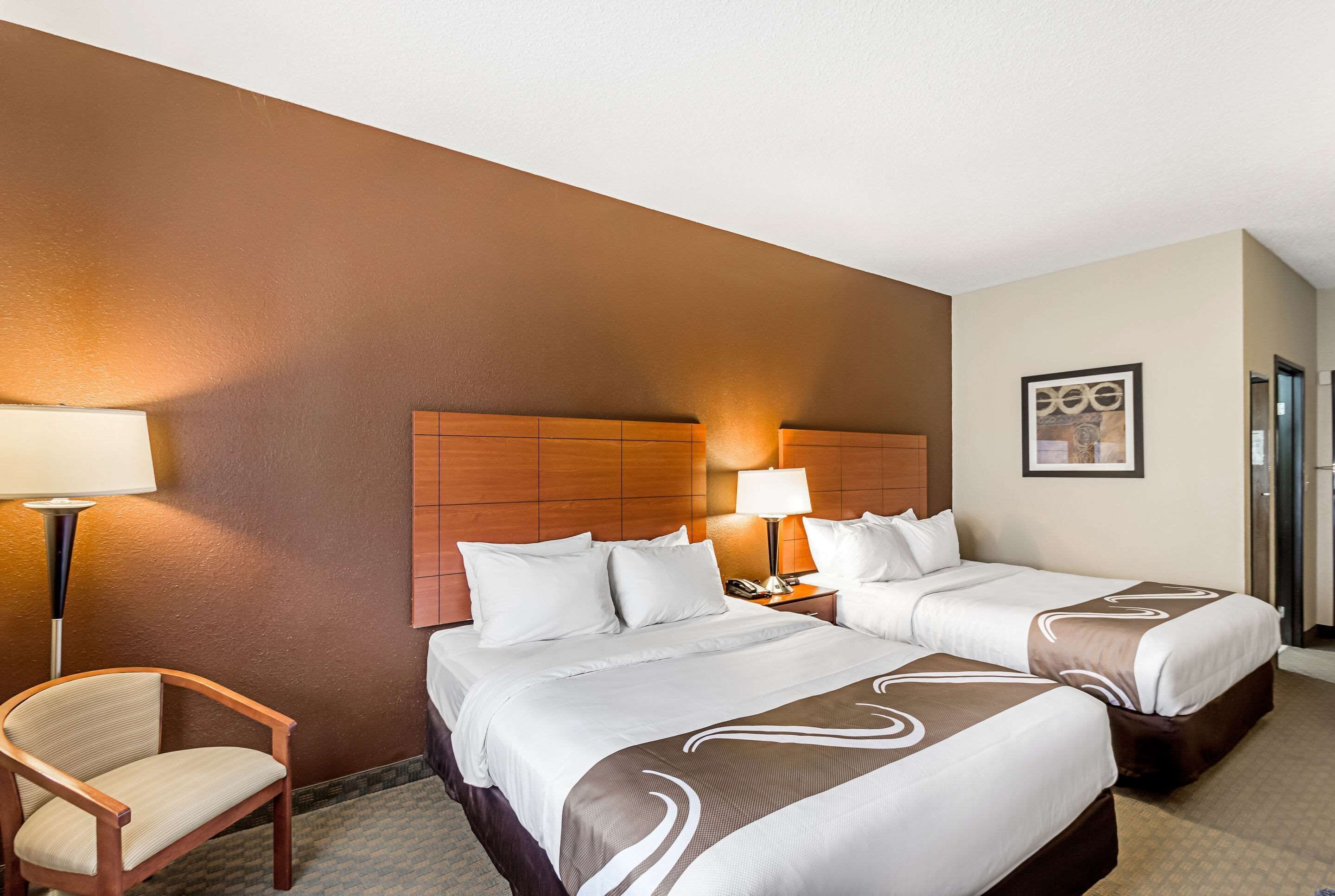 A hotel room with two beds, a wooden headboard, a chair, a lamp, and a framed picture on the wall.