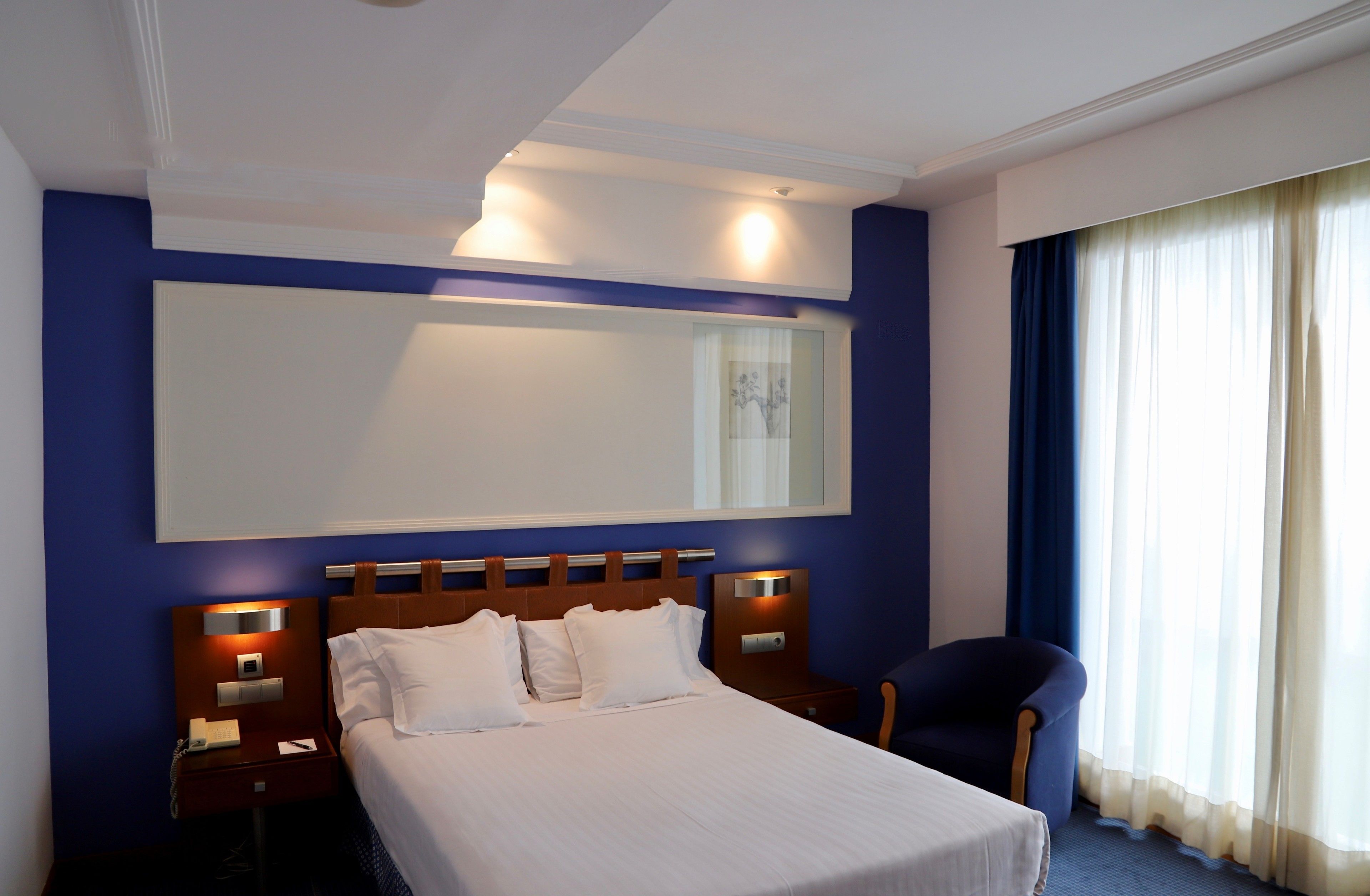 Habitación de hotel con una cama grande, una mesita de noche con lámpara, una silla y un espejo grande en la pared.
